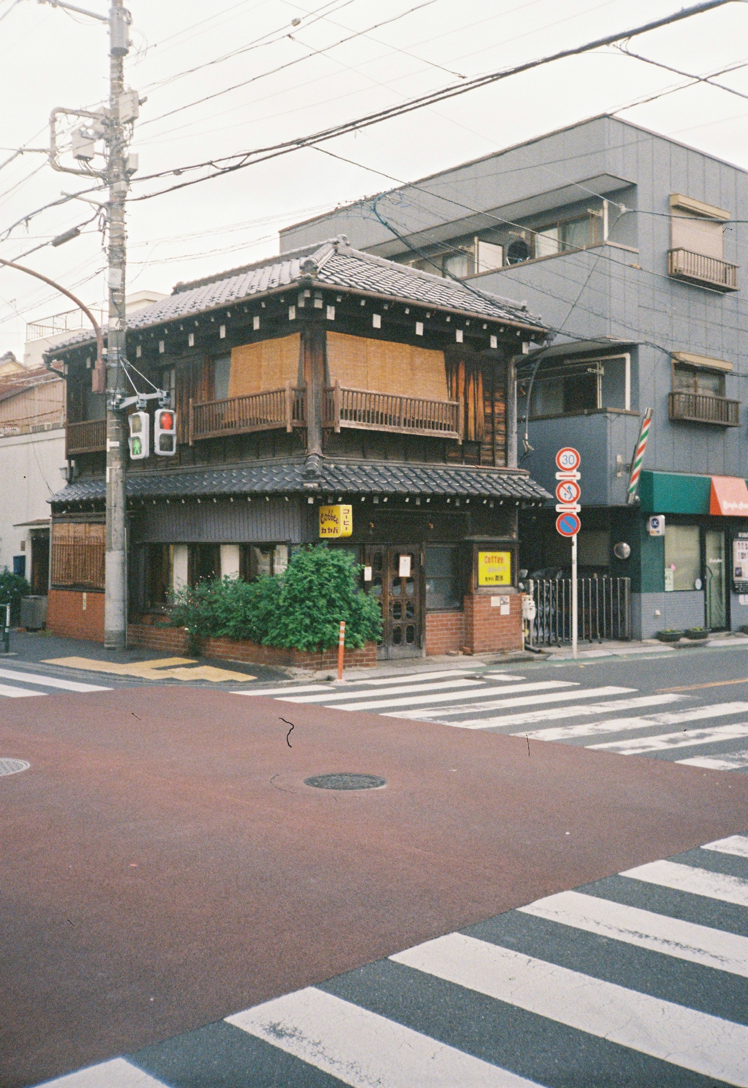 Edificio de estilo tradicional japonés junto a un edificio moderno, en una esquina de calle peatonal, con señales de tránsito y cruces peatonales.