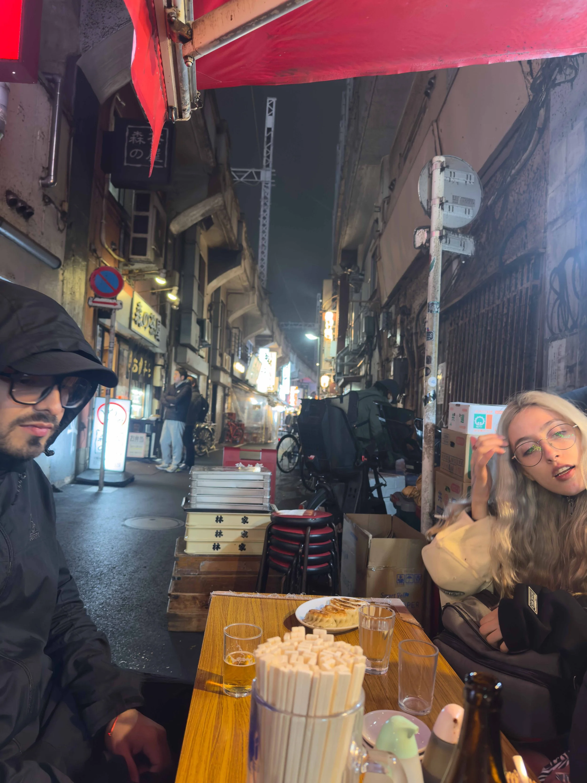 Personas en un restaurante al aire libre en la noche, sirviendo comida y bebidas en una mesa de madera, con fondo de calles iluminadas y gente caminando.