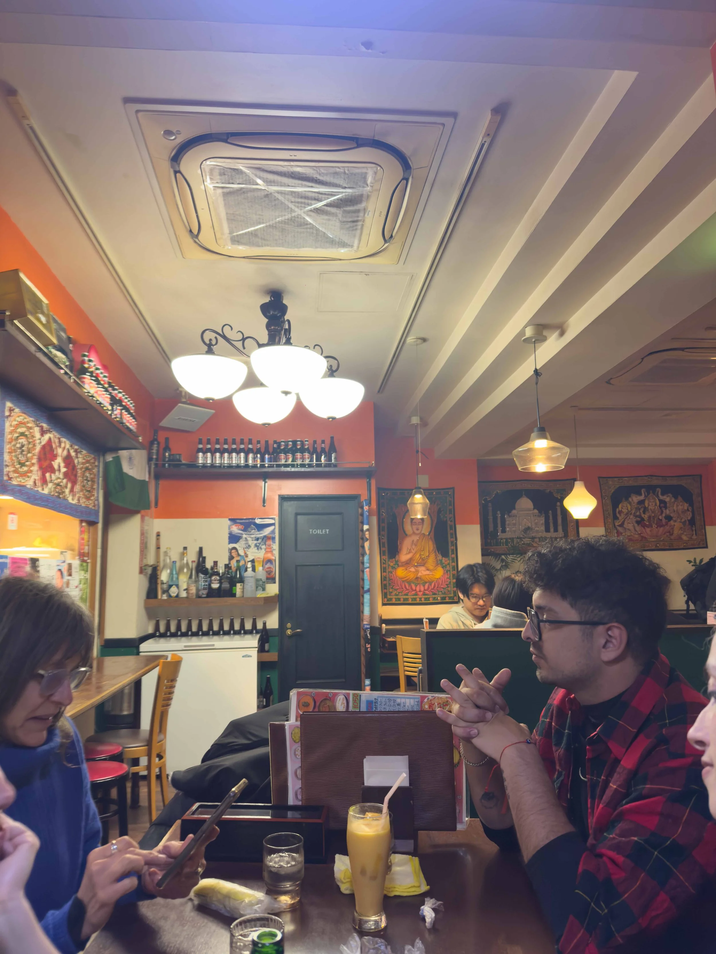 Personas sentadas en una mesa en un restaurante, con bebidas y comida, en un interior decorado con arte y elementos culturales, mientras conversan y usan teléfonos móviles.