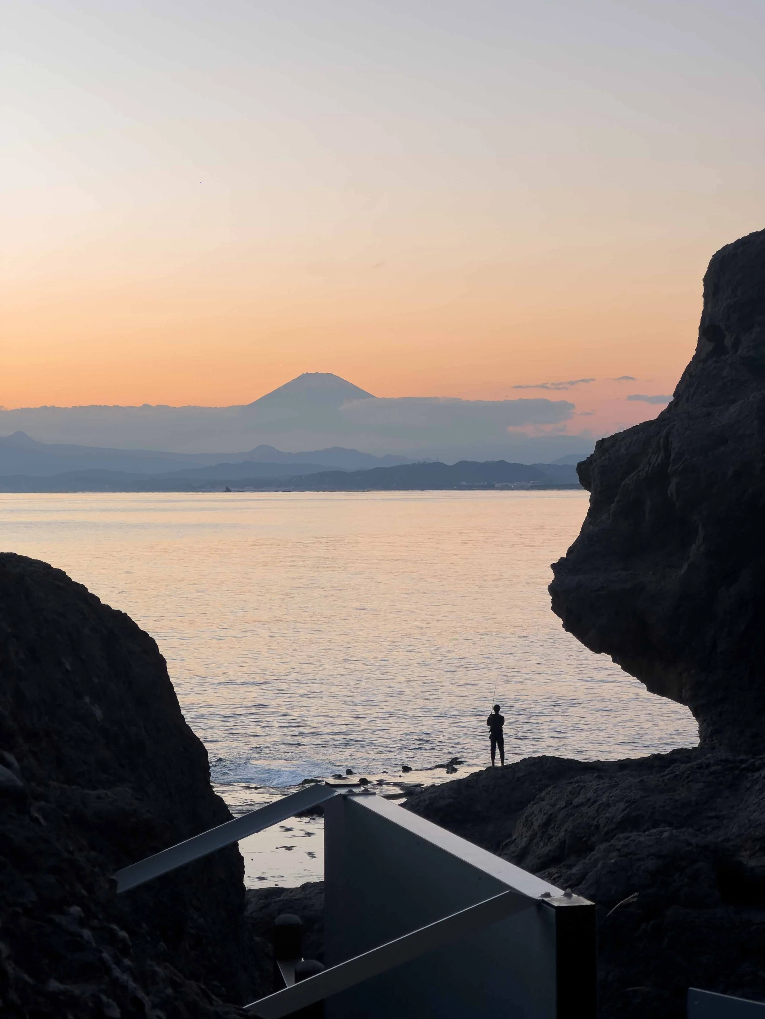 Paisaje costero en atardecer con vista de una montaña en el horizonte, un cielo con tonos suaves de naranja y gris, rocas en primer plano y una persona pescando en la orilla.