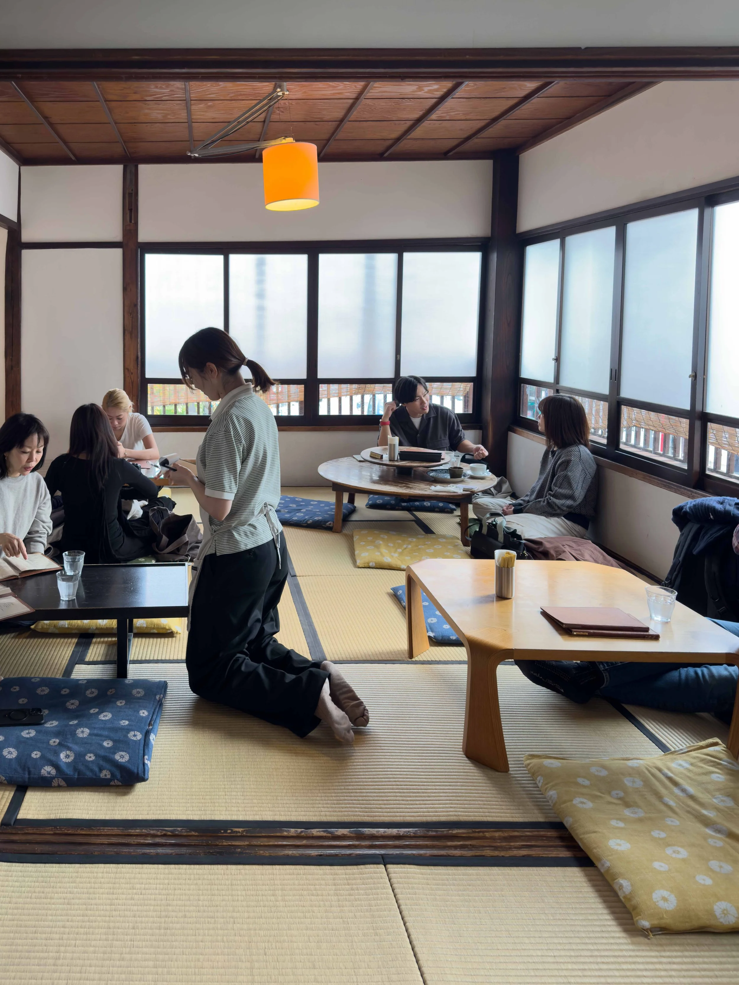 Varias personas en una sala de tatami, con mesitas bajas y cojines, algunas leyendo y otras conversando, con grandes ventanas y techo de madera.