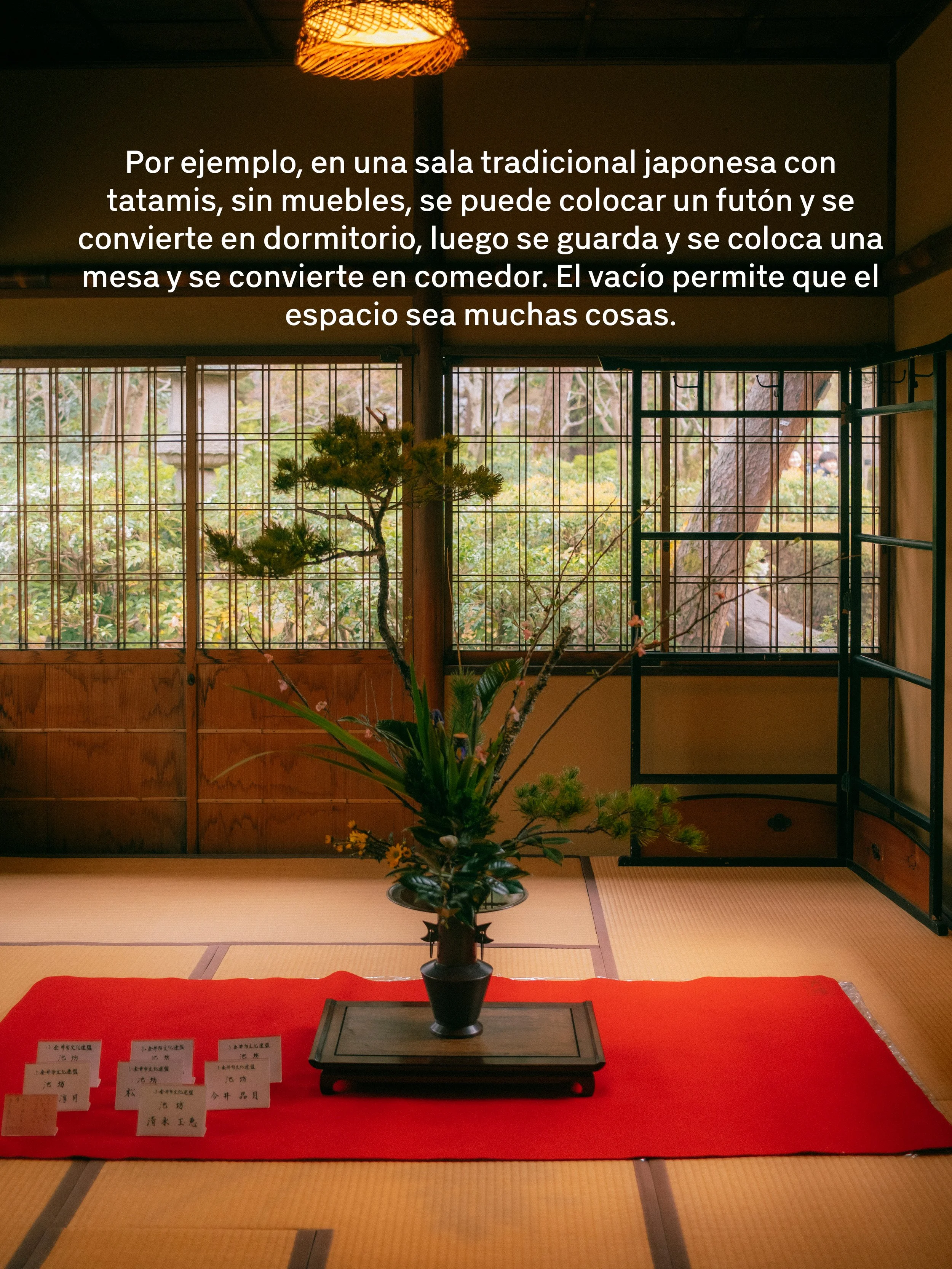 Una sala tradicional japonesa con tatamis, donde hay una mesa con un arreglo floral en un jarrón negro, en el fondo ventanas de madera y rejas, y un techo con lámpara de papel.