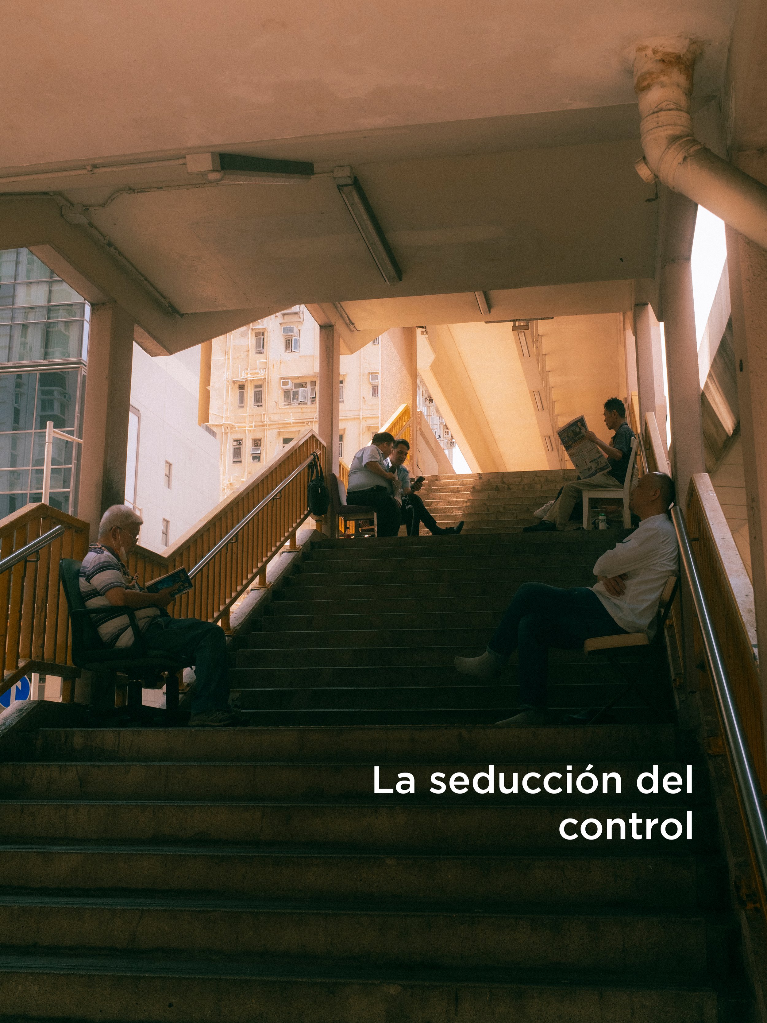 Personas sentadas en una banca en una escalera en un espacio abierto, leyendo, usando un teléfono y en conversaciones. El fondo muestra edificios altos y una estructura de techo con columnas.