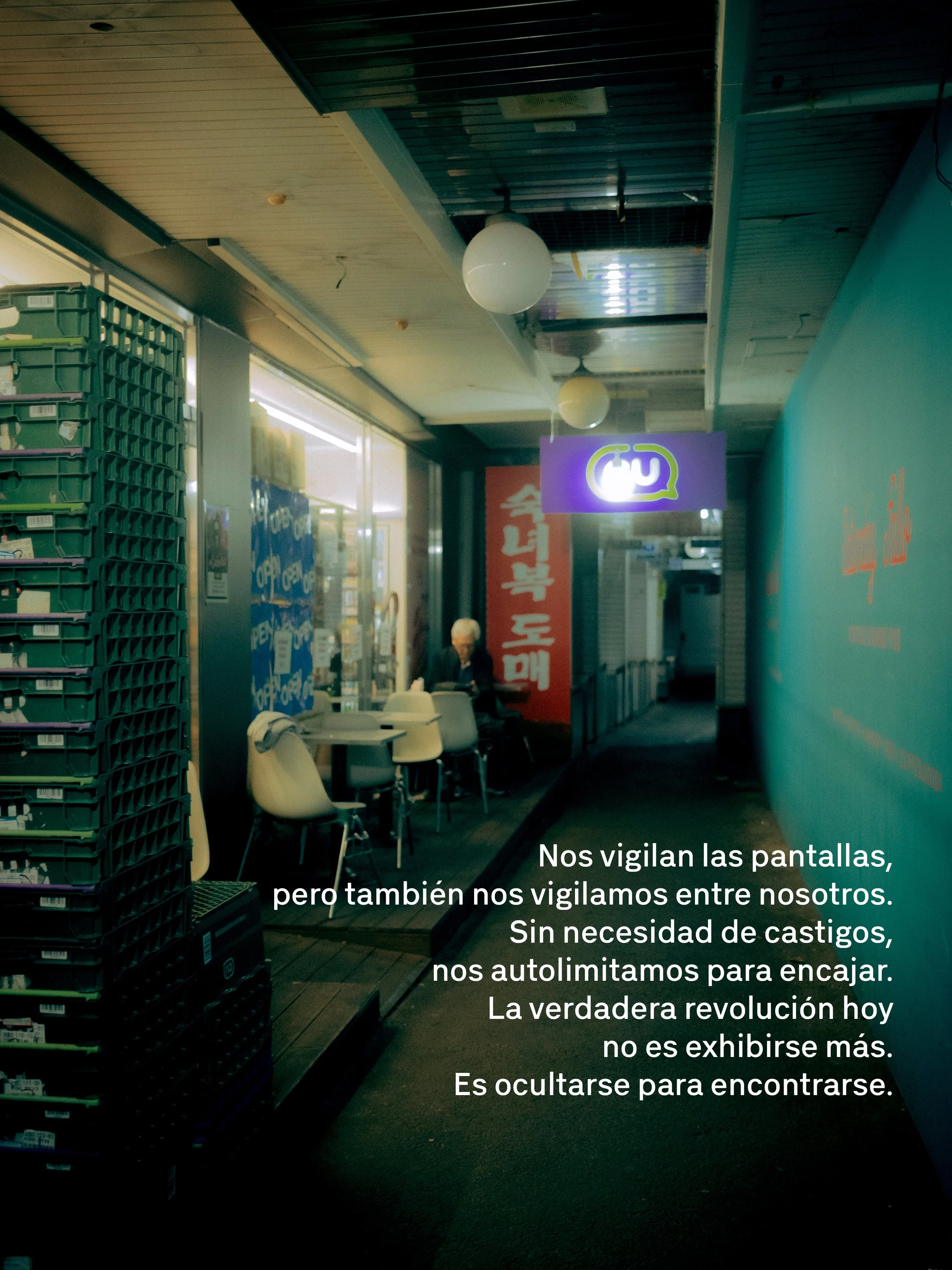 Imagen de un pasillo interior de un establecimiento comercial con mesas y sillas, carteles y contenedores de almacenamiento en la esquina izquierda. Sobre la imagen hay un texto en español que dice: "Nos vigilan las pantallas, pero también nos vigilamos entre nosotros. Sin necesidad de castigos, nos autolimitamos para encajar. La verdadera revolución hoy no es exhibirse más. Es ocultarse para encontrarse."