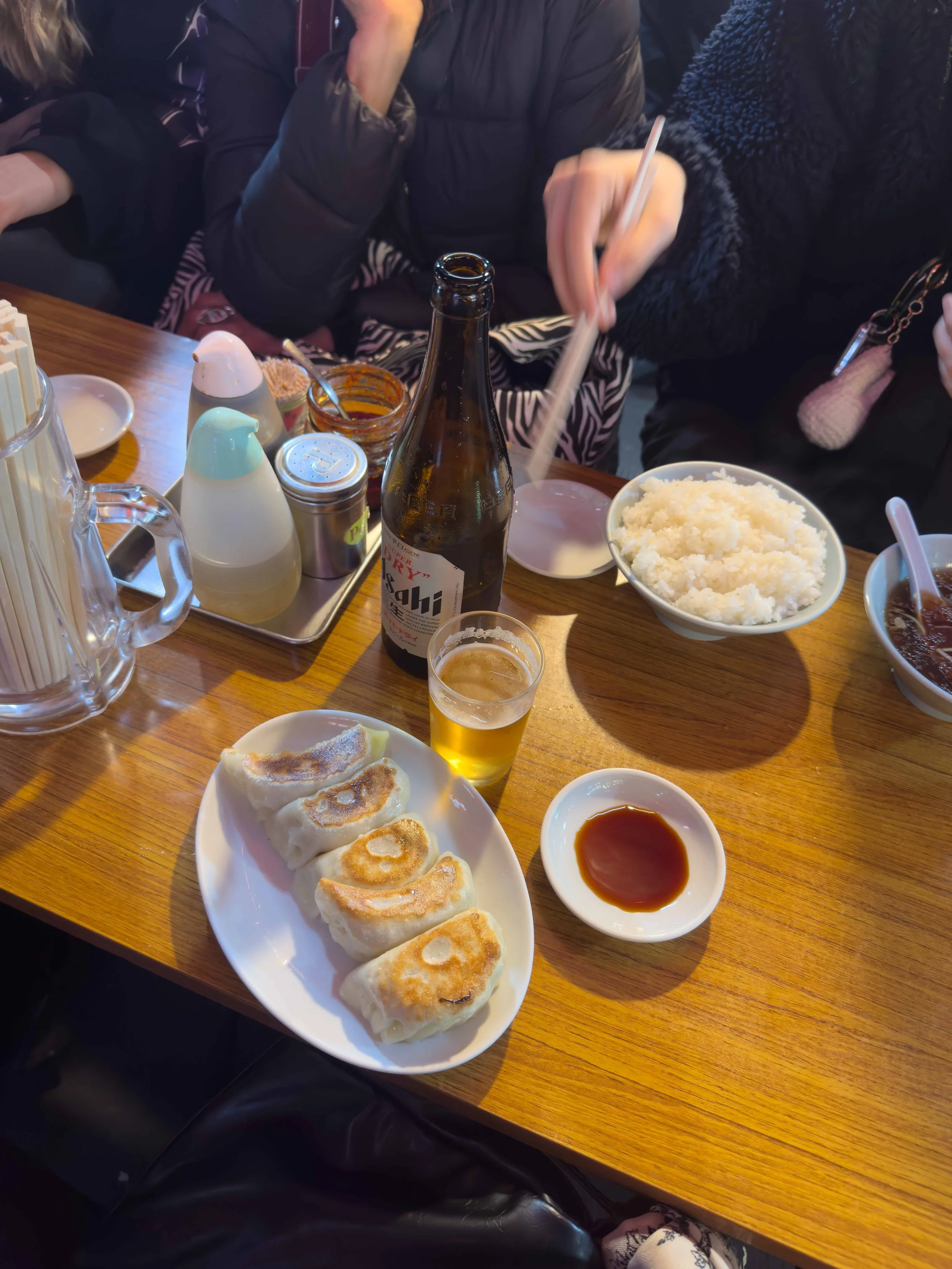 Plato con cinco empanadas doradas, acompañadas de salsa en un pequeño plato, en una mesa de restaurante con otras comidas y bebidas, incluyendo un plato de arroz y una botella de cerveza.
