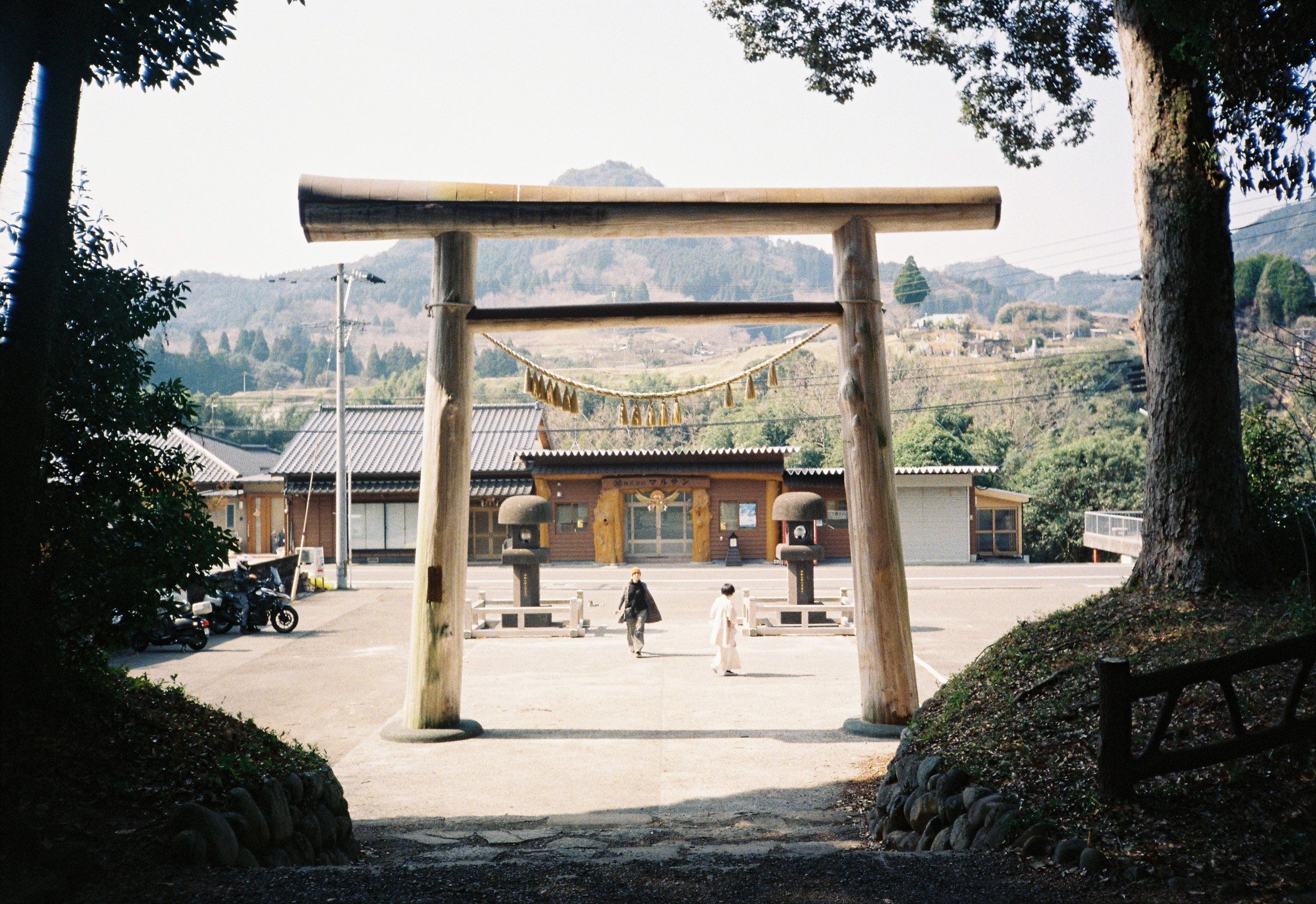 Un arco tradicional de entrada a un templo shinto en un entorno rural, rodeado de árboles y con montañas a lo lejos, en Japón.