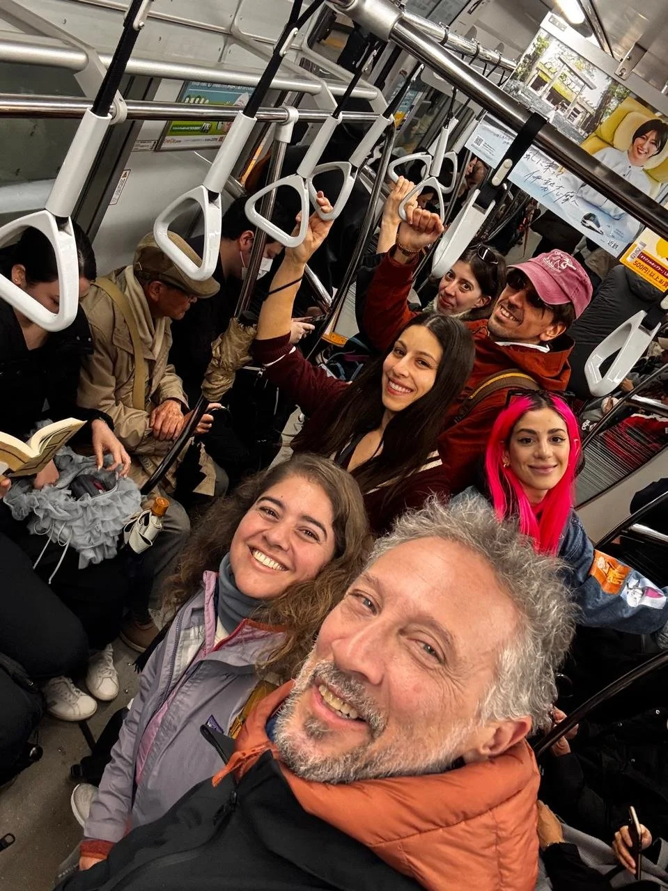 Grupo de personas en un autobús sentado, algunos mirando sus teléfonos y otros sonriendo o levantando la mano. La escena parece ser una selfie tomada por uno de los pasajeros.