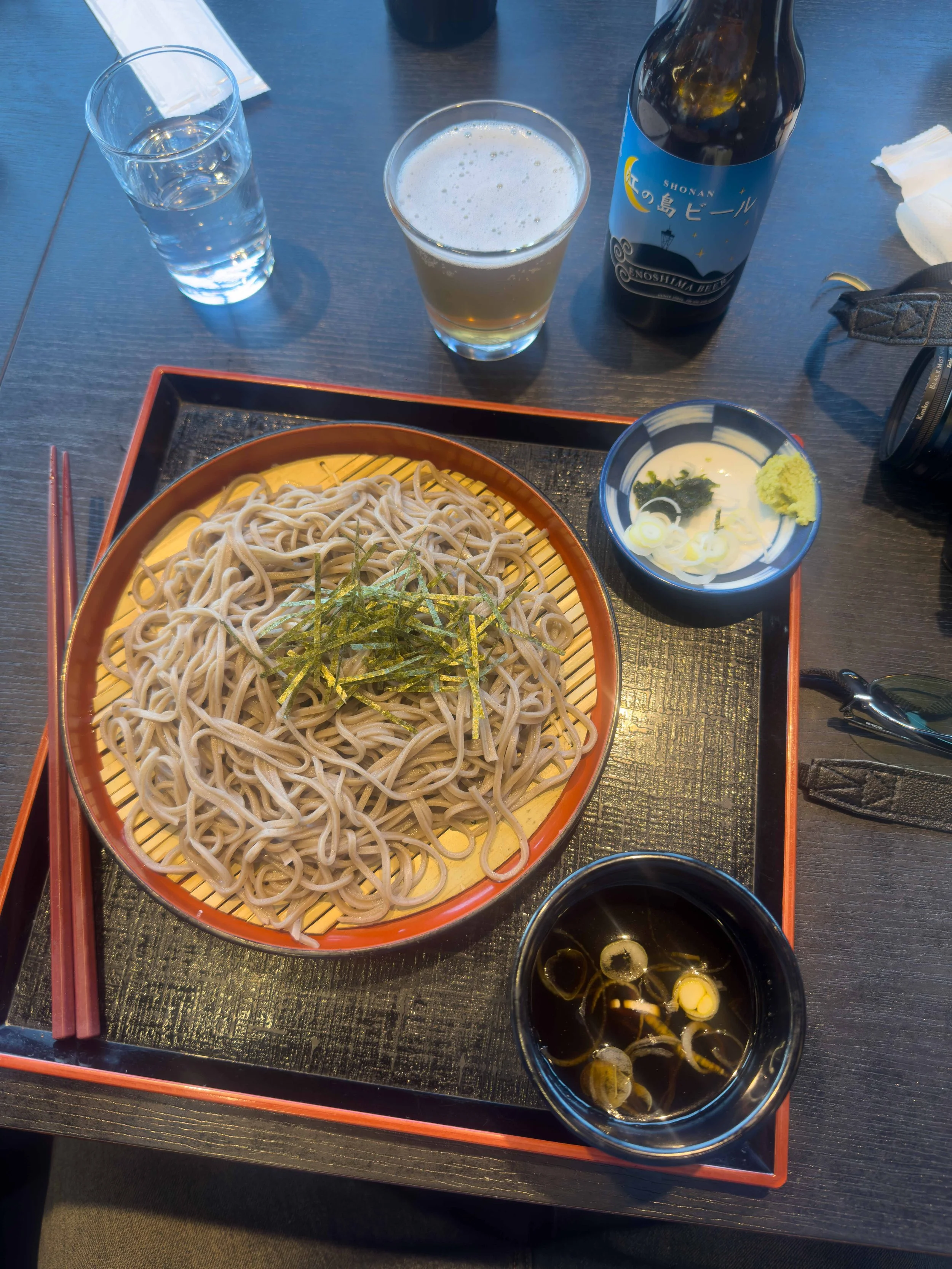 Plato de fideos soba con tiras de alga y un plato pequeño con crema, cebolla y wasabi, junto a una taza de sopa con cebolla en rodajas y unas botellas de cerveza y agua en una mesa de madera.