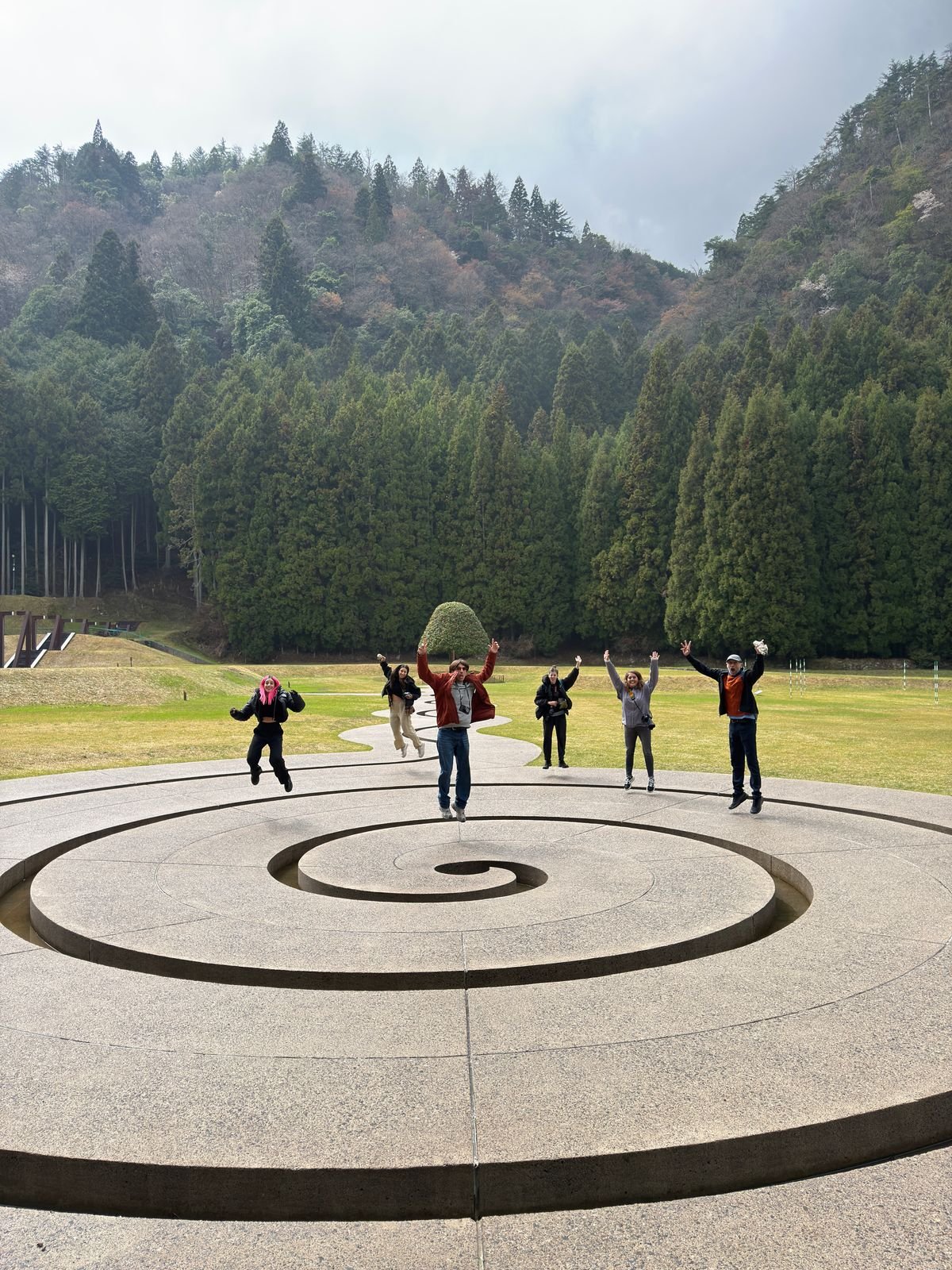 Varias personas brincando y posando sobre una escultura circular en un parque con árboles y montañas de fondo.