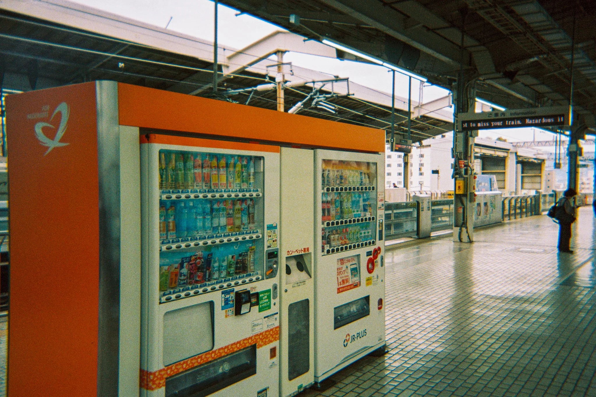 Estación de tren con máquinas expendedoras de refrescos y comida, y pantalla digital con información de los trenes en japonés, en un espacio cubierto y con piso de baldosas.