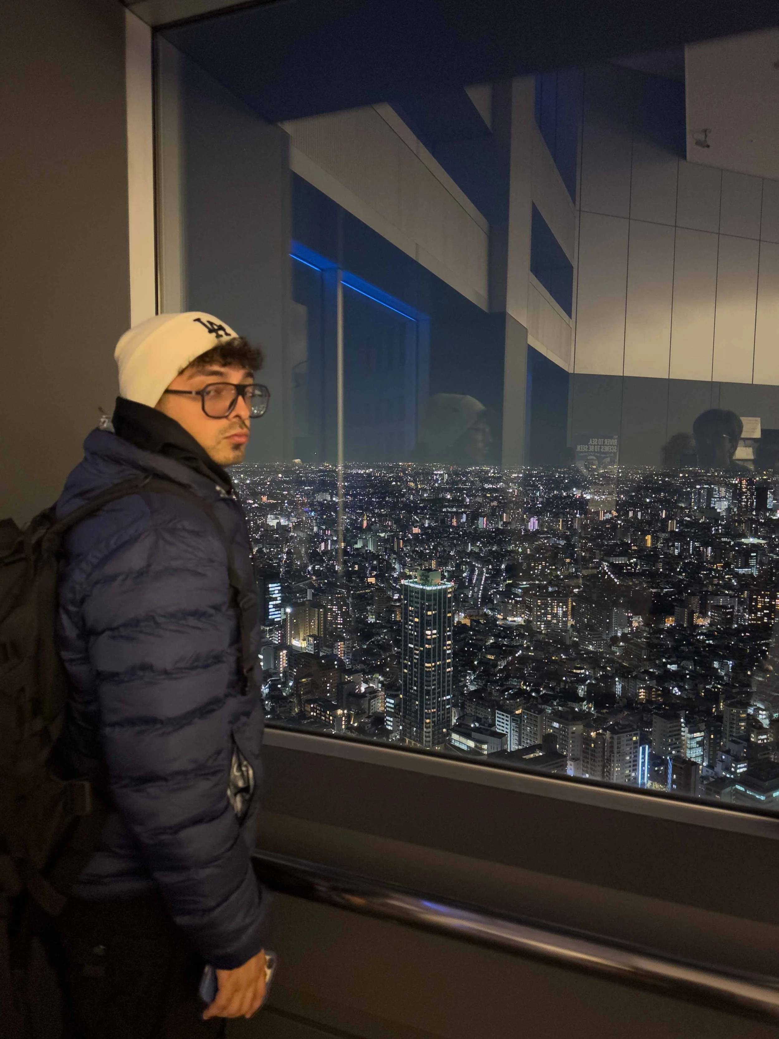 Joven con gorra, lentes y chaqueta oscura, en un elevador con vista nocturna de la ciudad