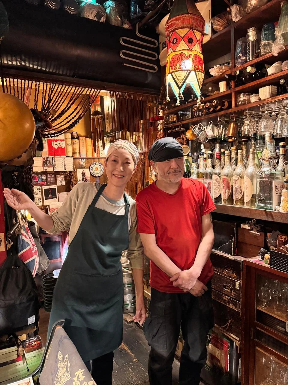 Dos personas, una mujer con delantal y un hombre con gorro, en un ambiente de bar o restaurante con botellas y decoraciones culturales.