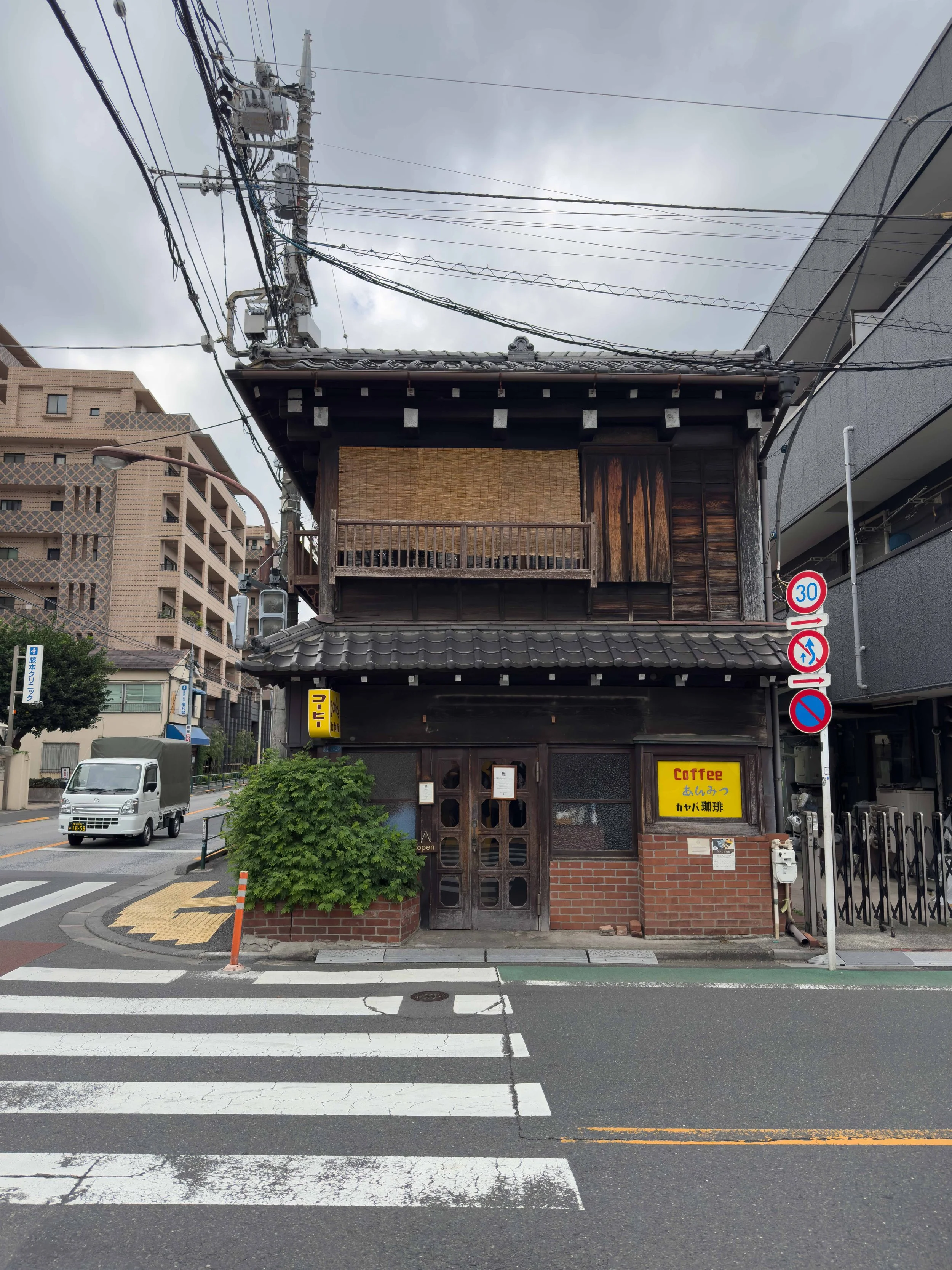 Edificio de estilo tradicional japonés en una calle urbana, con un cartel que indica café y señal de límite de velocidad de 30 km/h. El cielo está nublado y hay vehículos y semáforos en la calle.