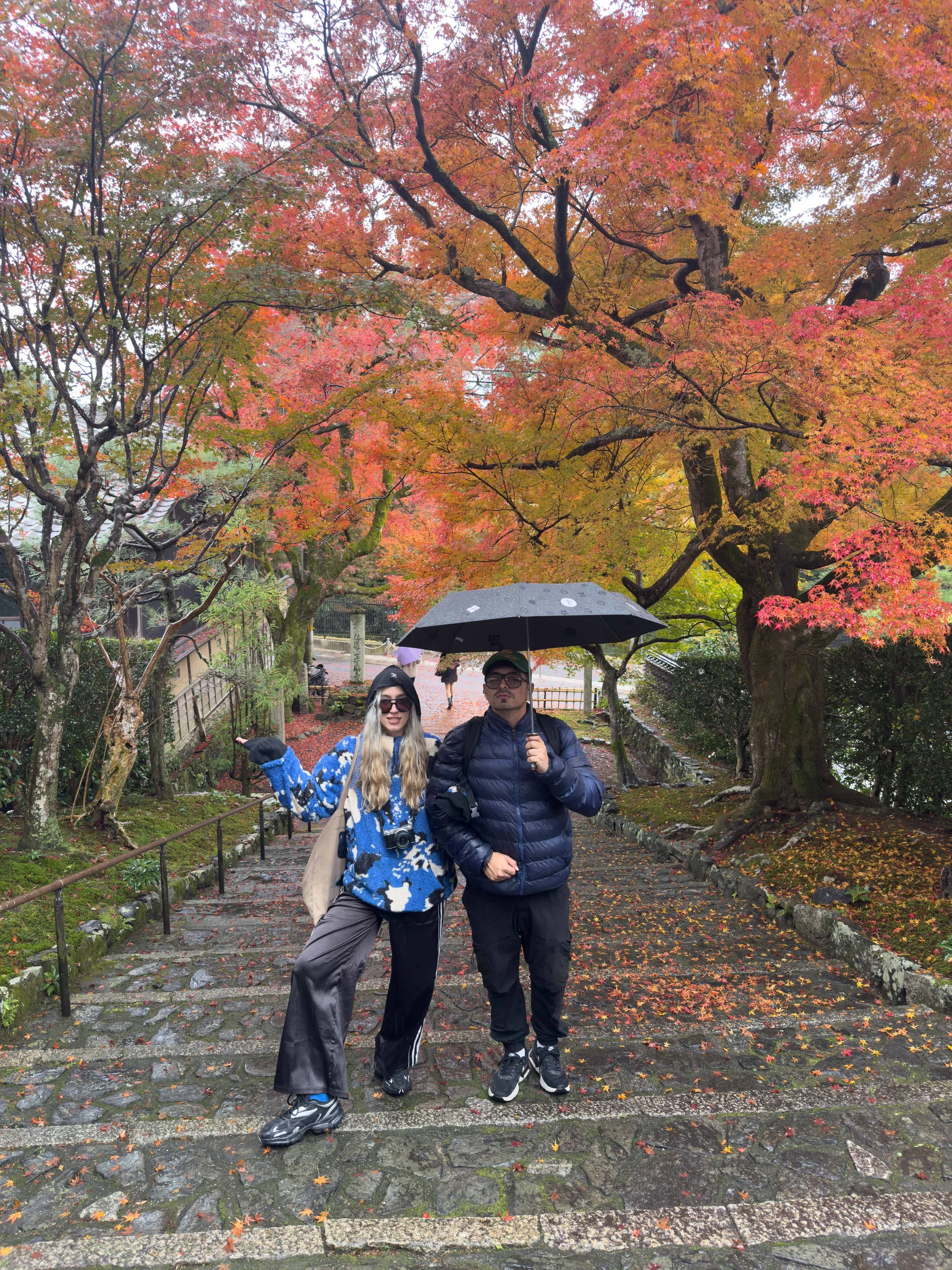 Dos personas con paraguas en un sendero, rodeadas de árboles con hojas de otoño en tonos anaranjados, rojos y amarillos, con una ligera lluvia.