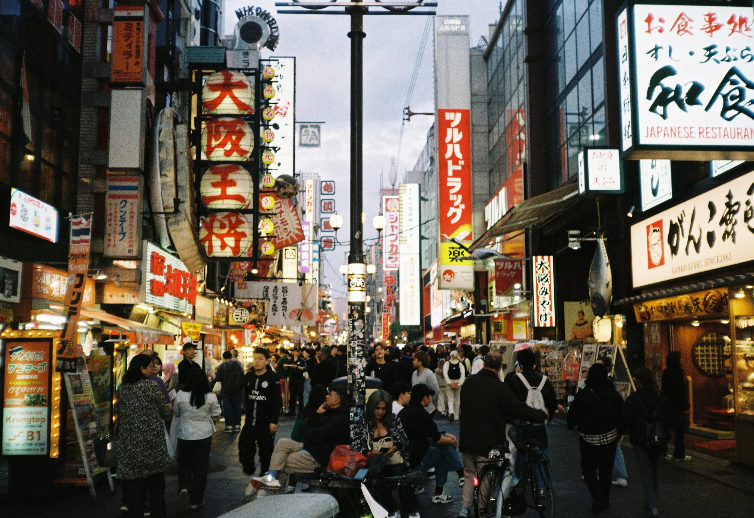 Una calle llena de gente con numerosos letreros luminosos en japonés y tradiciones comerciales japonesas, en un entorno urbano concurrido.