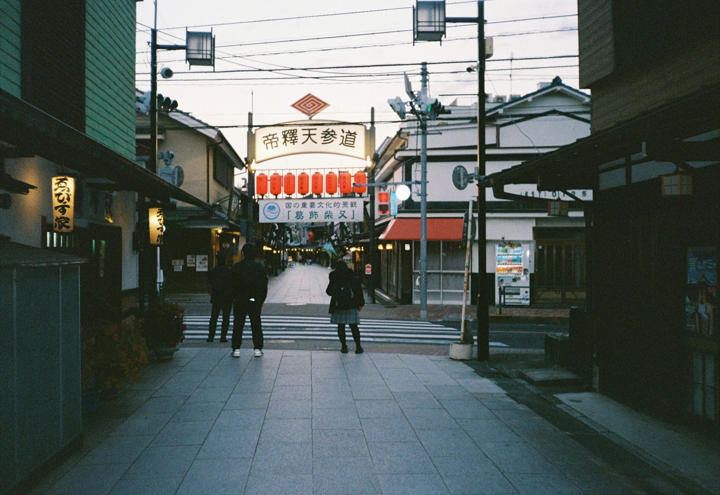 Calle peatonal en un barrio comercial japonés durante el atardecer con letreros en japonés, linternas rojas y personas caminando.