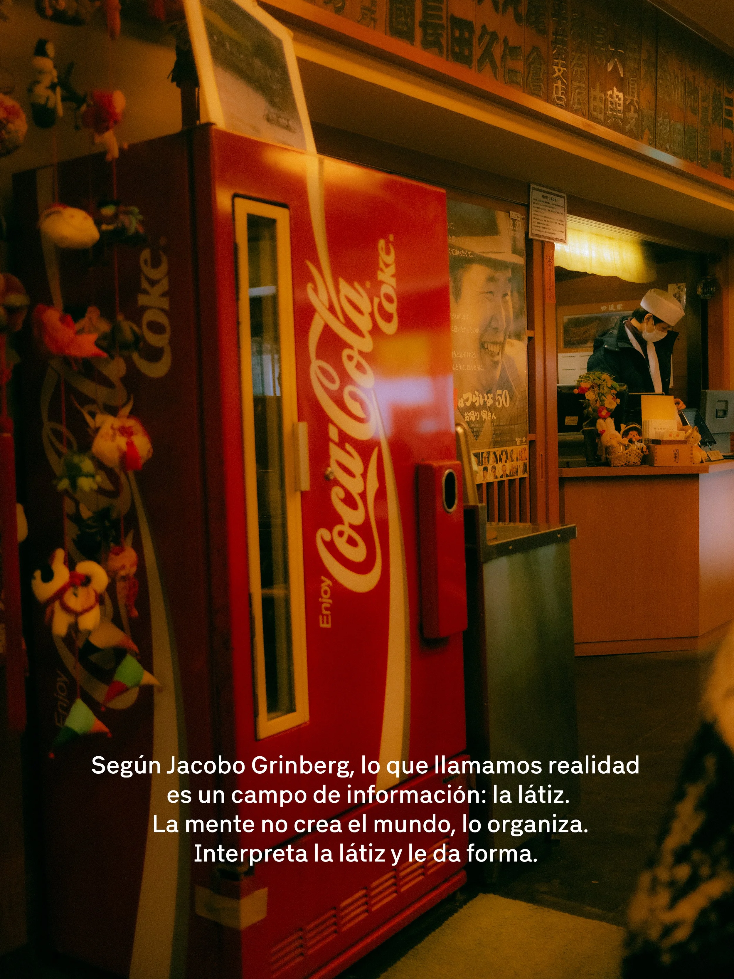 Una máquina expendedora de bebidas Coca-Cola en un establecimiento, acompañada de un cartel y decoraciones con juguetes colgantes a la izquierda. En el fondo, un empleado con uniforme, máscara y gorro, atendiendo en un mostrador.
