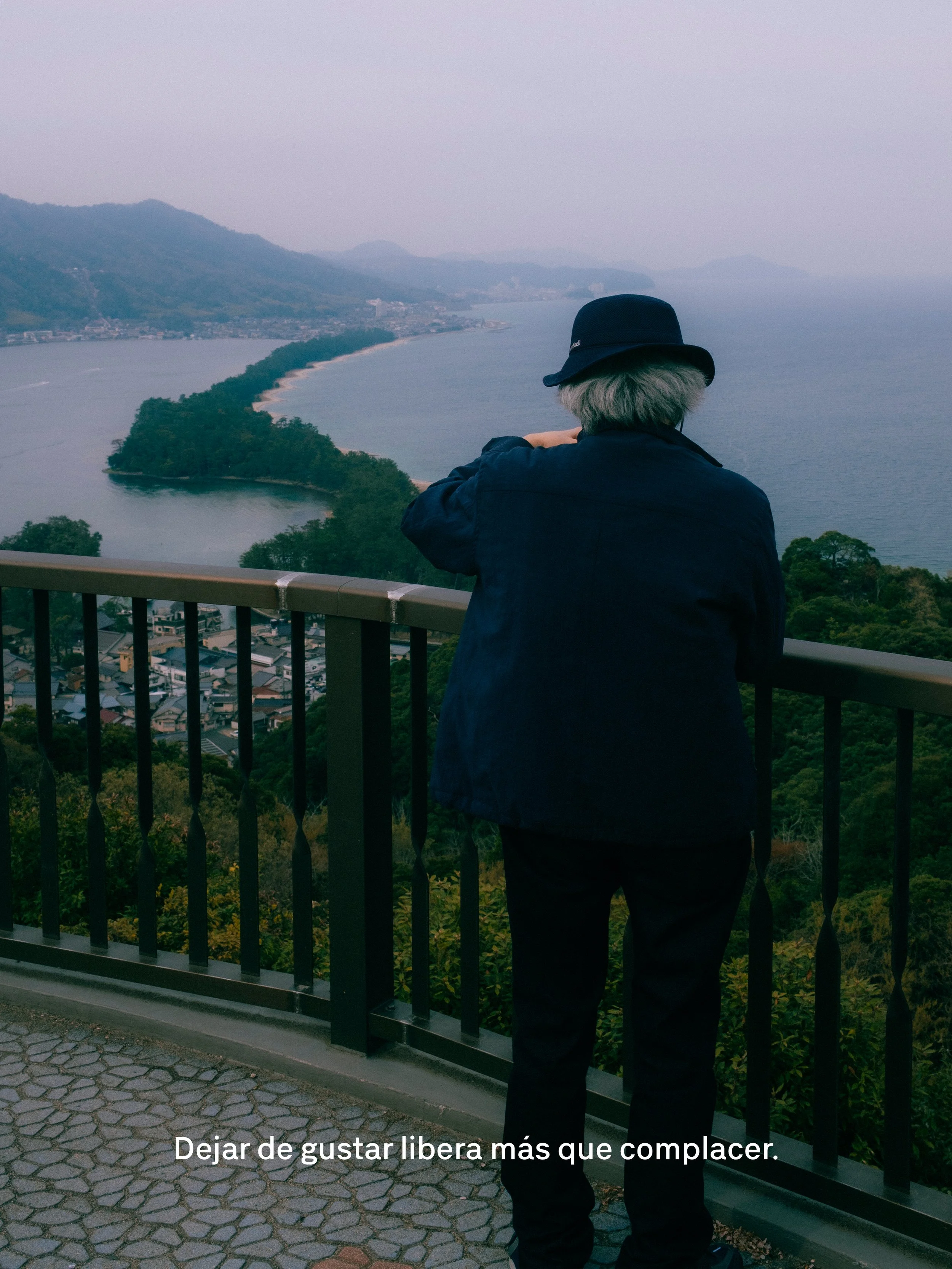 Persona adulta de espaldas, con sombrero negro, observando un paisaje de río con colinas y vegetación desde un mirador.