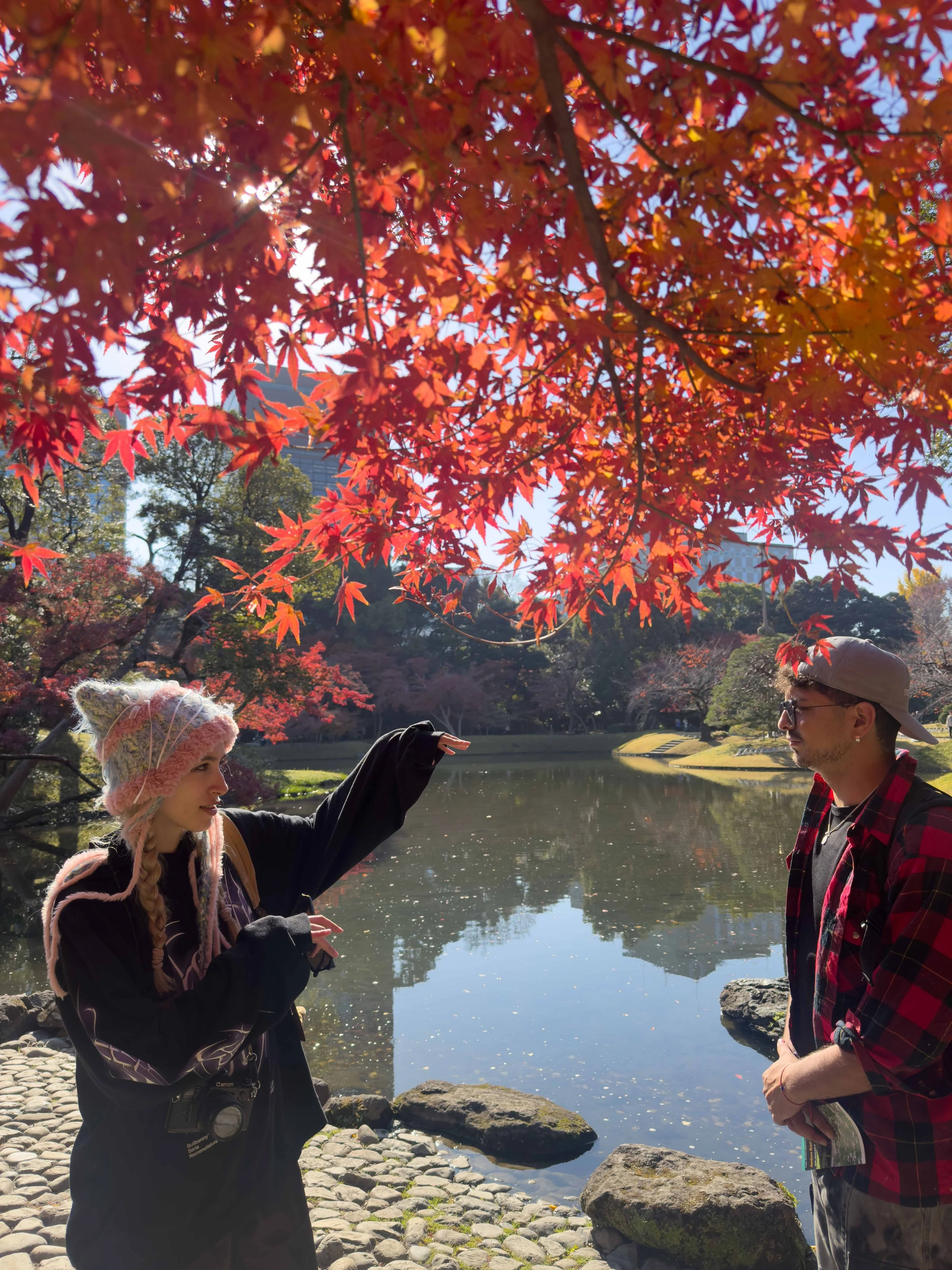 Dos personas junto a un río con árboles de hojas rojas en otoño. Una mujer con gorro y cámara, un hombre con gafas y gorra, en un parque.