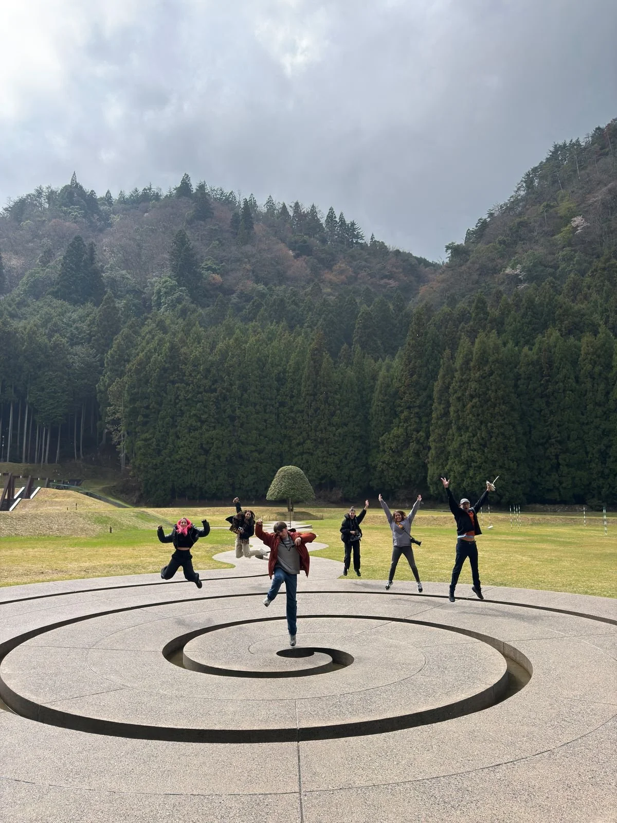 Grupo de seis personas brincando y posando en un parque con fondo de montañas y árboles, en un día nublado.