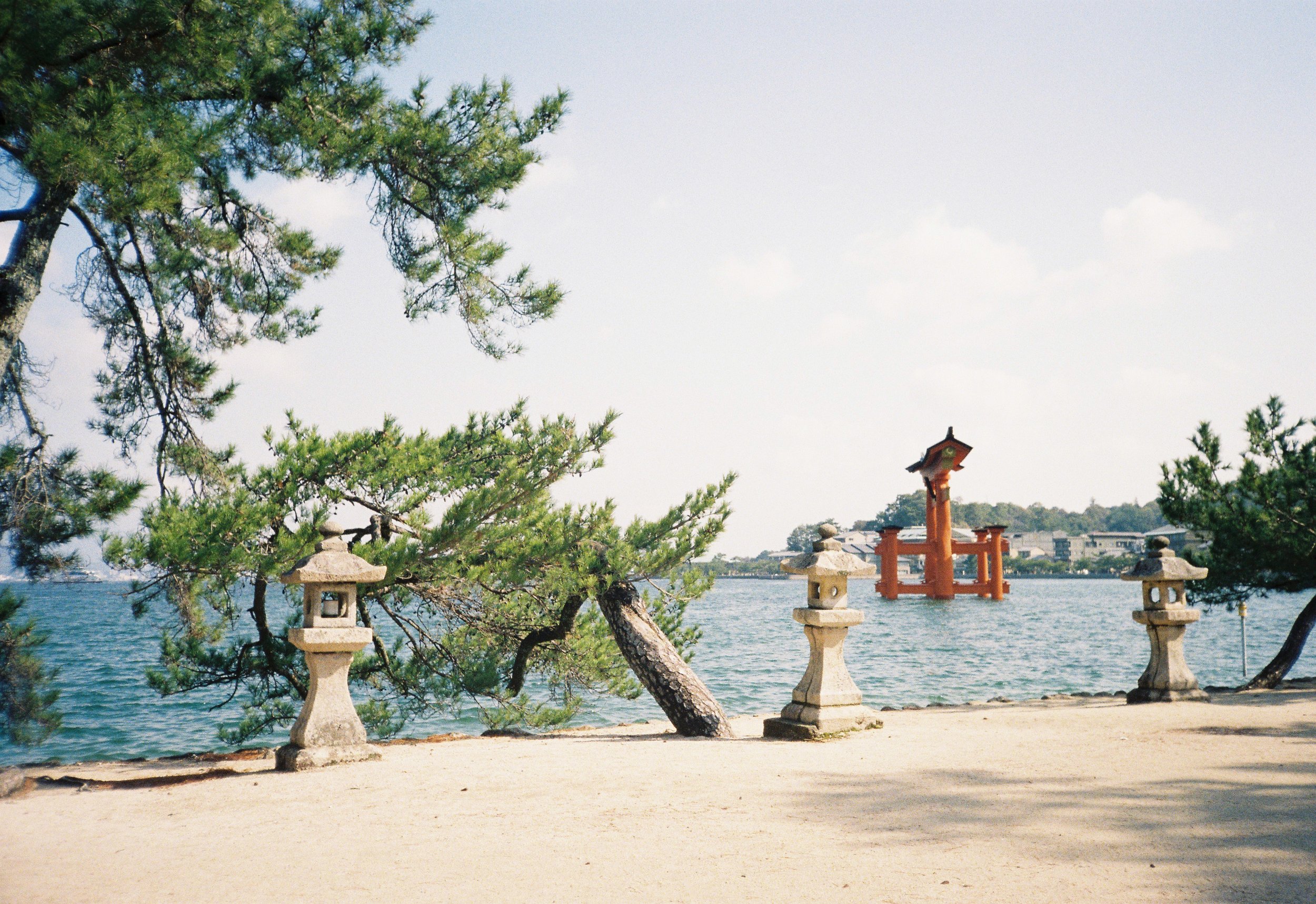 Vista de un lugar junto al río con árboles de pino, linternas tradicionales de piedra, un torii rojo frente al agua, y viviendas al fondo bajo un cielo parcialmente nublado.