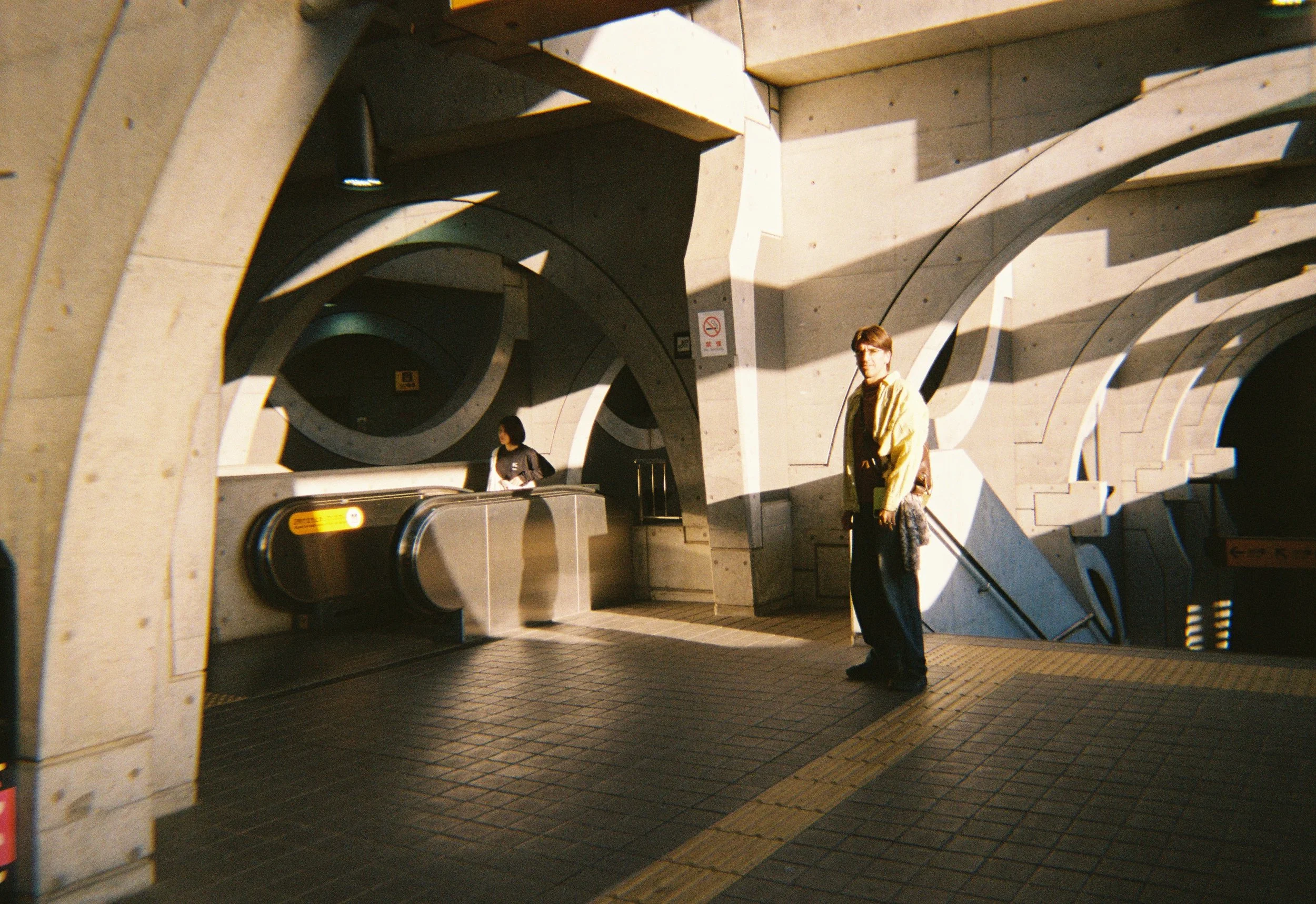 Persona con gafas y chaqueta amarilla en un ingreso de estación de tren, con arquitectura moderna de concreto y escaleras eléctricas.