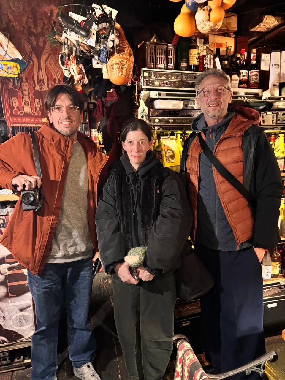 Tres personas sonrientes posando en una tienda con estantes llenos de bebidas y objetos diversos, rodeadas de artículos decorativos y bolsas.