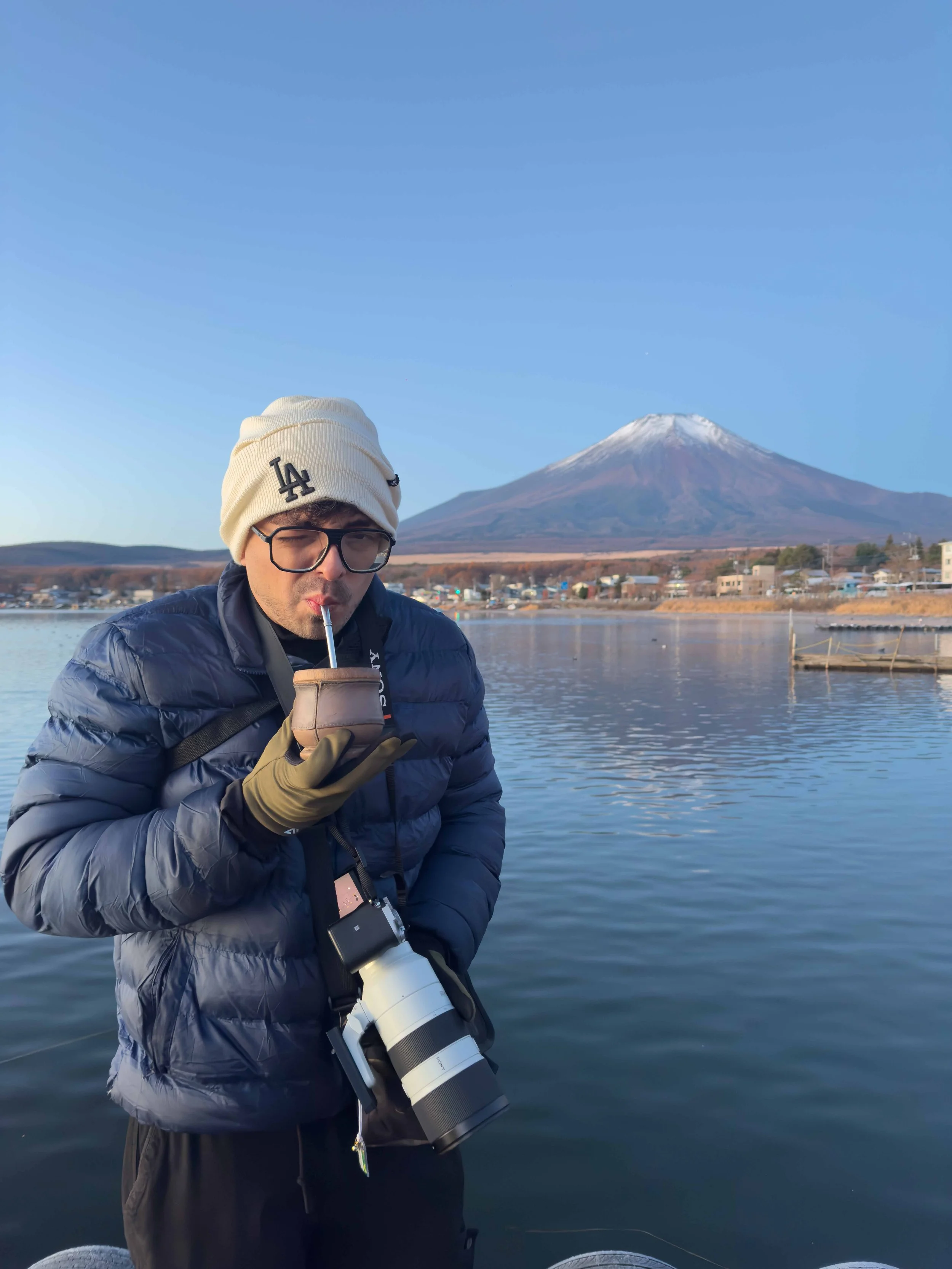 Hombre vestido con abrigo y gorro, sosteniendo una cámara telescópica junto a un cuerpo de agua con una montaña nevada al fondo.
