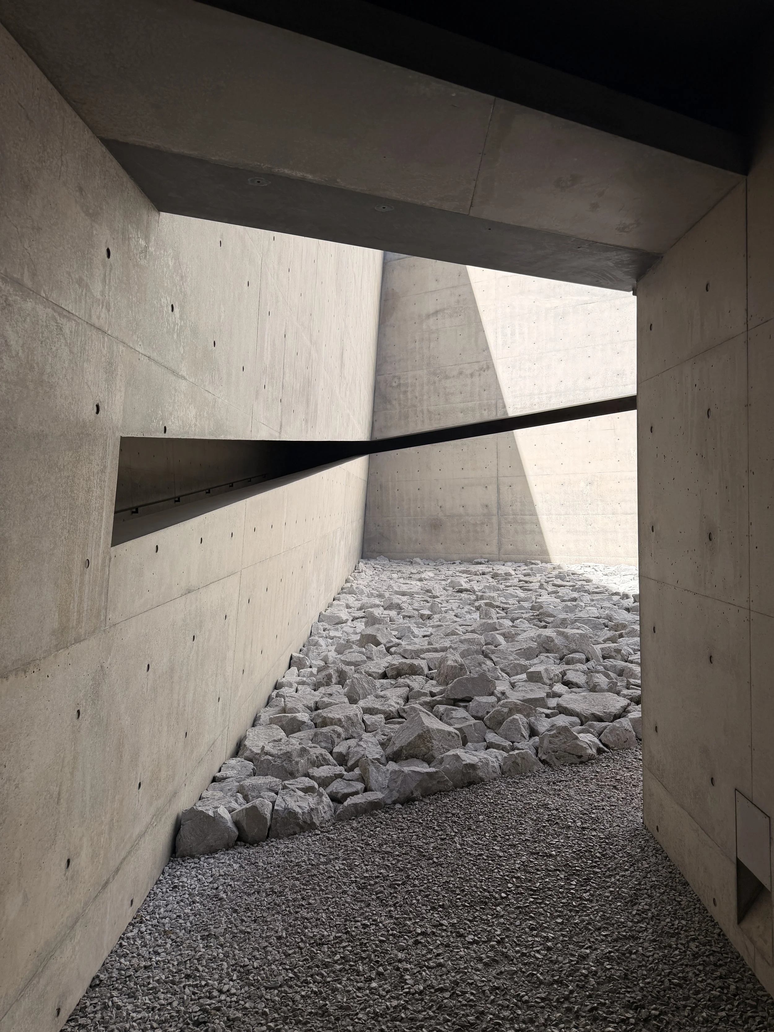Pasillo o espacio abierto con paredes de concreto, suelo cubierto de rocas y piedras, con luz natural entrando desde arriba.