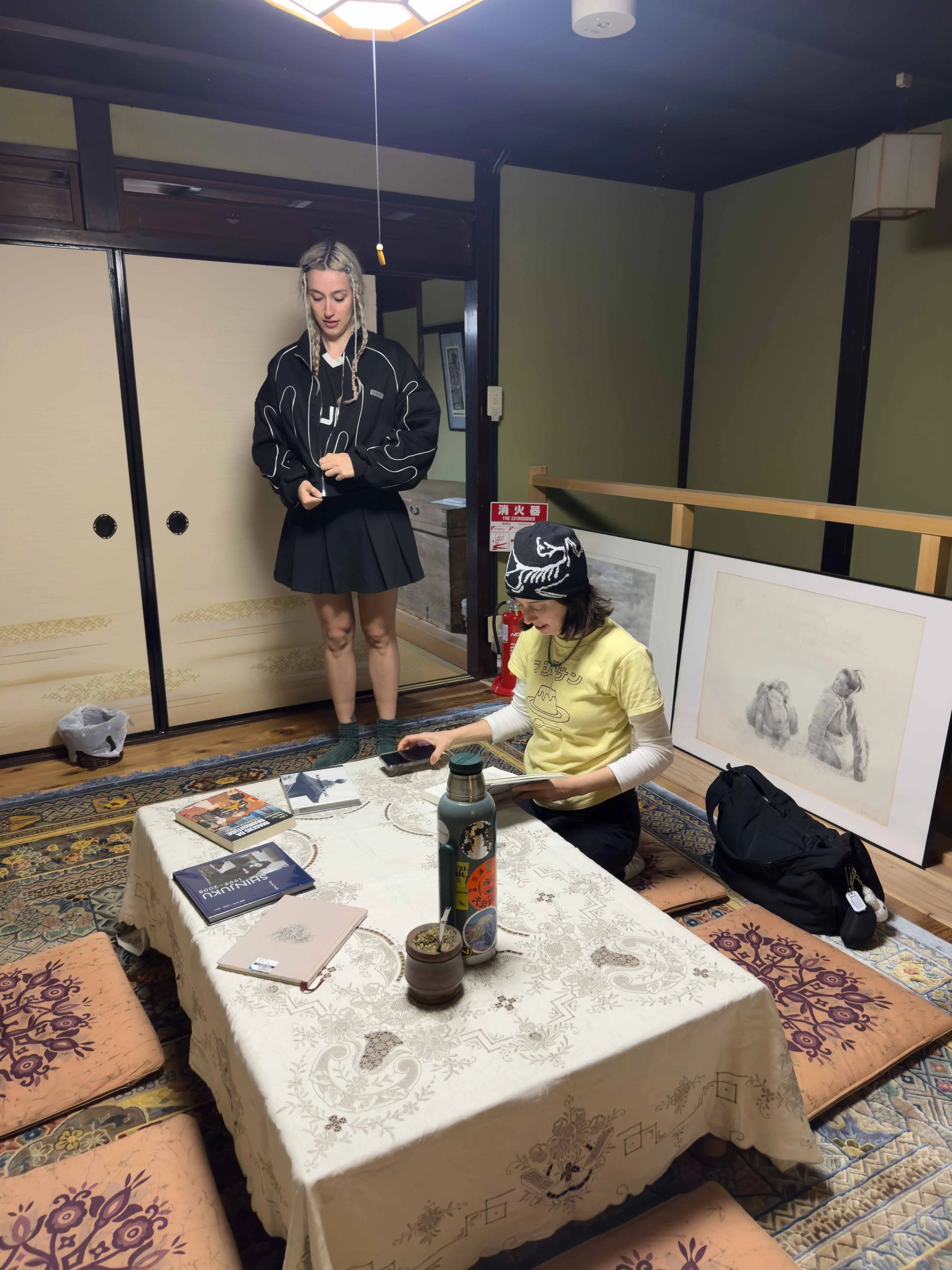 Una habitación con paredes de color verde y puertas correderas de estilo tradicional japonés. En el centro hay una mesa cubierta con un mantel blanco, rodeada de cojines para sentarse. Sobre la mesa hay libros, una planta en maceta, una botella de ag