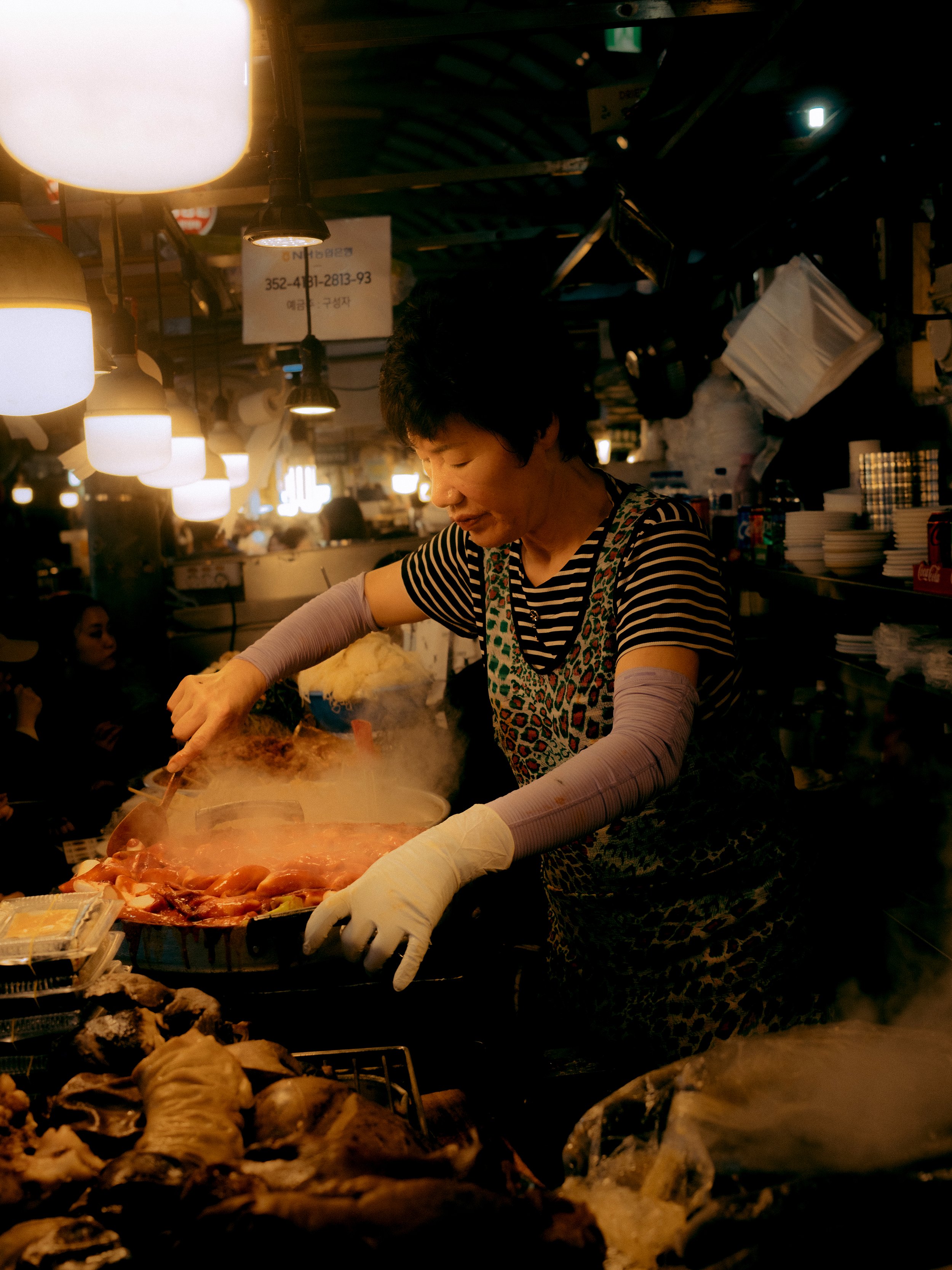 Una mujer preparando carne en un puesto de comida en un mercado o feria, con muchas luces y otros puestos en el fondo.