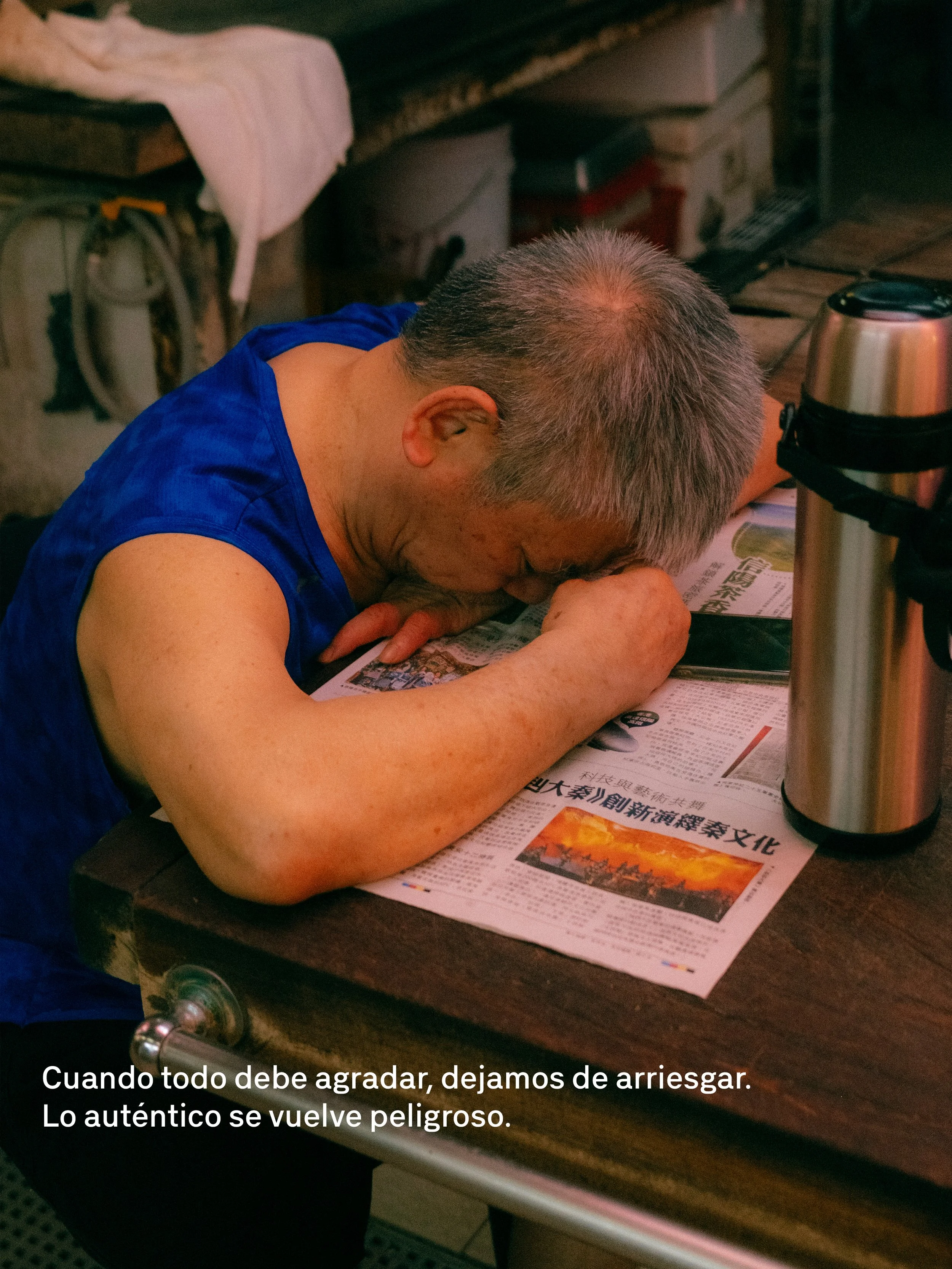 Hombre con cabello canoso, vestido con camiseta azul, apoyado sobre una mesa leyendo un periódico en japonés. En la mesa hay una botella térmica y otros objetos. Comentario en la imagen: "Cuando todo debe agradar, dejamos de arriesgar. Lo auténtico se vuelve peligroso."