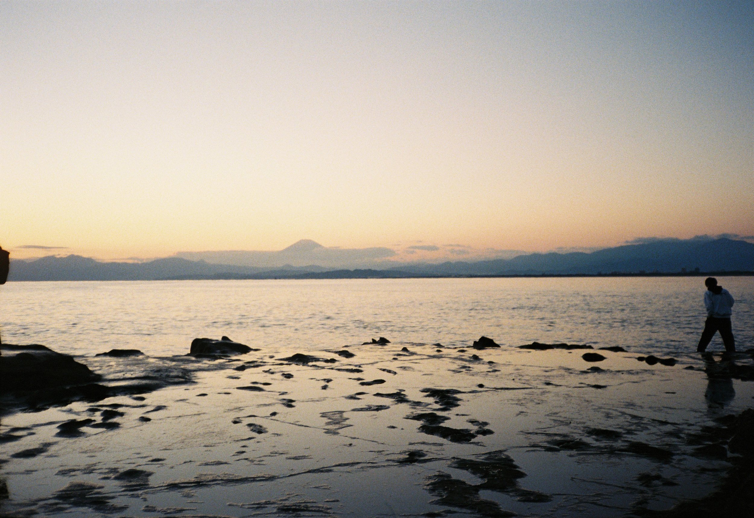 Persona caminando cerca de las rocas en la orilla del mar durante el atardecer, con montañas al fondo y cielo despejado.