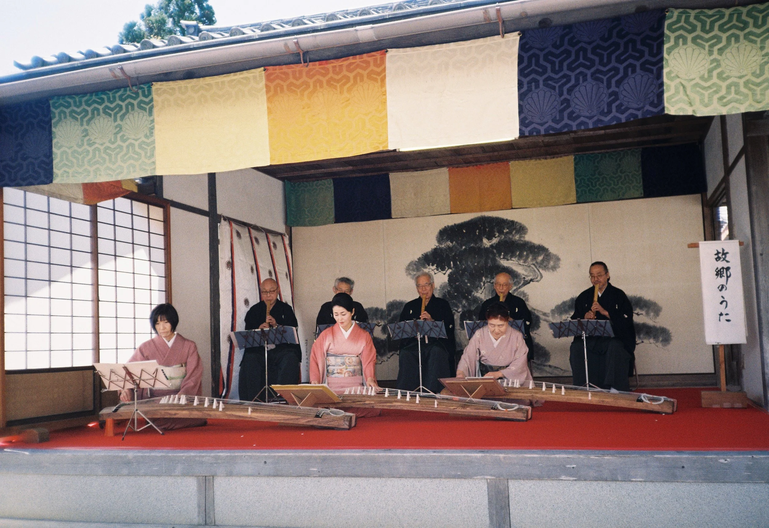 Un grupo de músicos tocando instrumentos tradicionales japoneses en un escenario decorado con banners y una pintura de un árbol.