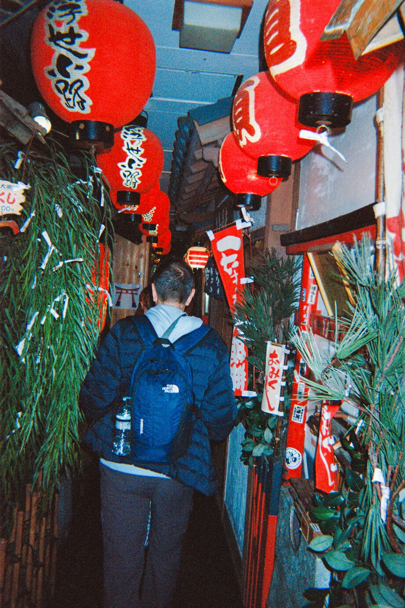 Una persona con mochila de senderismo camina por un pasillo decorado con faroles rojos con caracteres japoneses, banderines y plantas decorativas a los lados.
