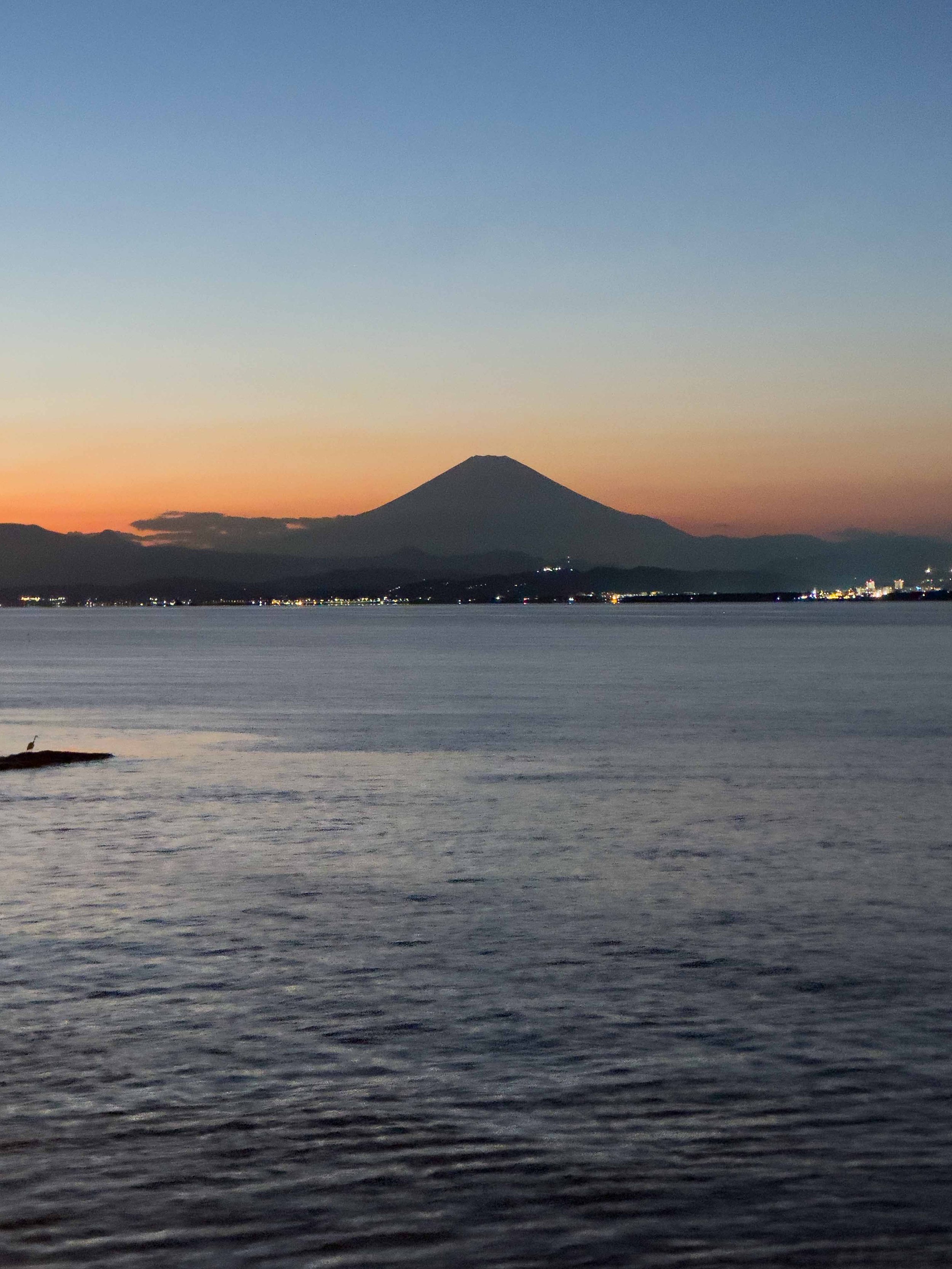 Atardecer sobre el mar con vista del volcán conico en el fondo y luces de la ciudad en la orilla.