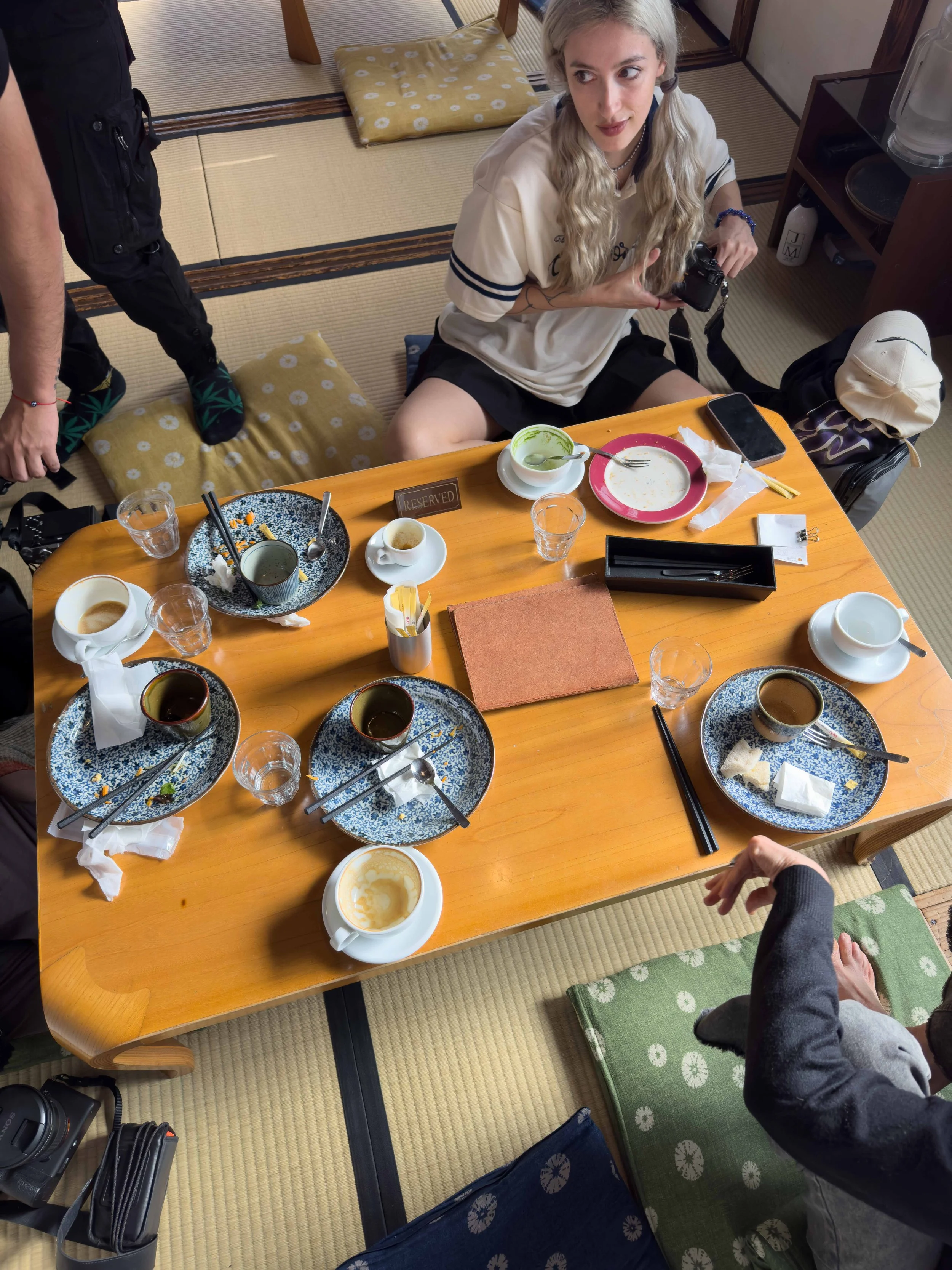 Una mujer con cabello rubio y corto, sentada en el suelo de un comedor de estilo japonés, rodeada de platos vacíos y una taza con restos de comida. Ella sostiene una cámara en las manos y mira hacia arriba. Hay una persona con pantalones negros y cal