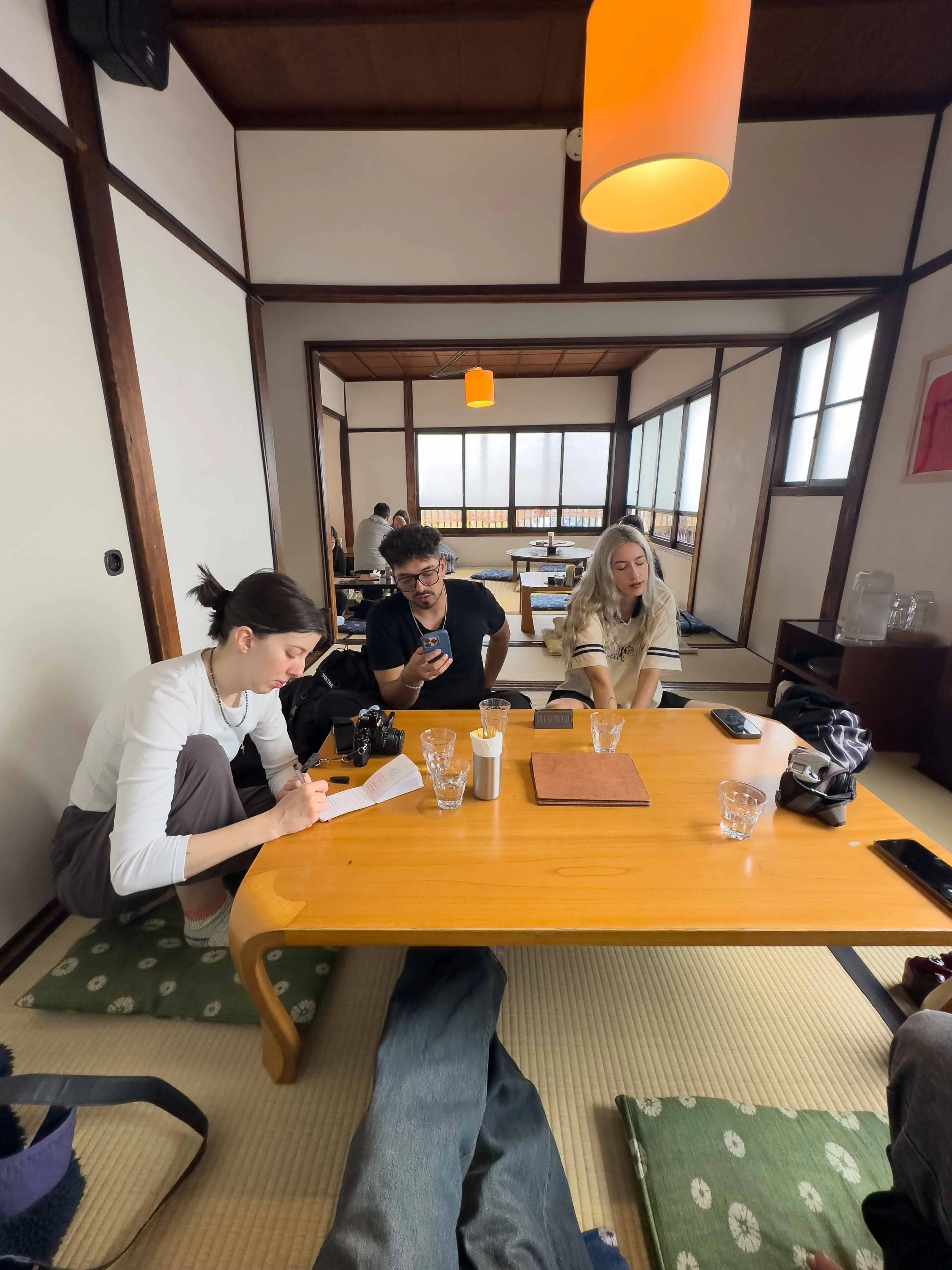 Grupo de personas sentadas en una mesa de madera en una restaurant japonés, algunos mirando sus teléfonos y otros leyendo un libro. La habitación tiene paredes blancas, ventanas grandes y un suelo de tatami.