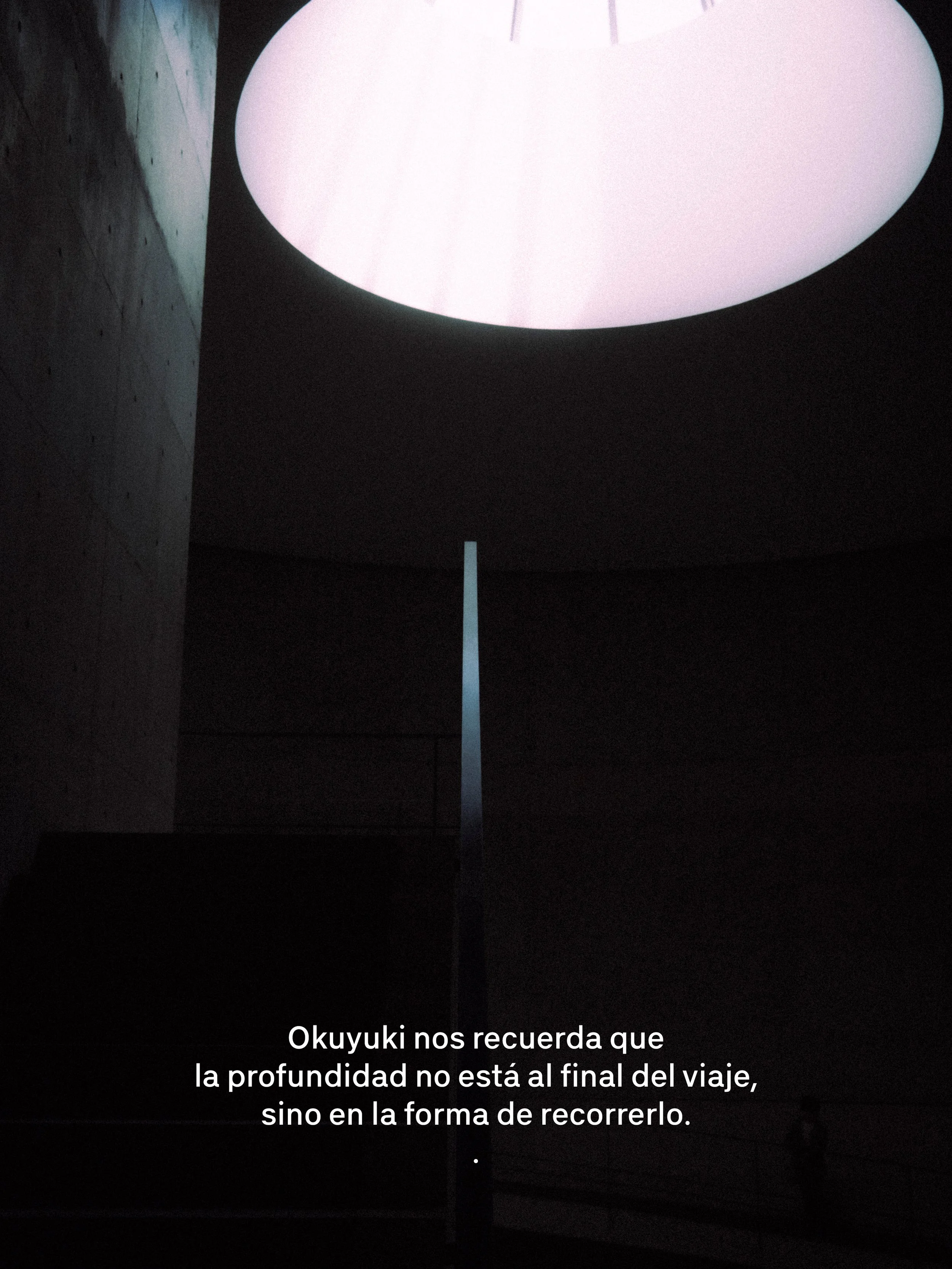 Una estructura moderna con una luz circular en el techo y una columna delgada en el centro, con fondo oscuro y una pared de cemento a la izquierda.