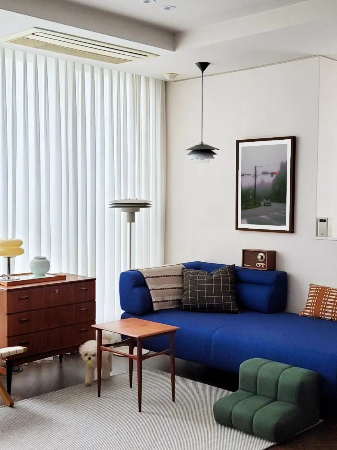 Sala de estar con sofá azul, mesa de madera, cuadros y lámparas de pie vintage, cortinas blancas largas y piso de madera con alfombra clara. Un perro pequeño de color blanco se asoma debajo de la mesa.