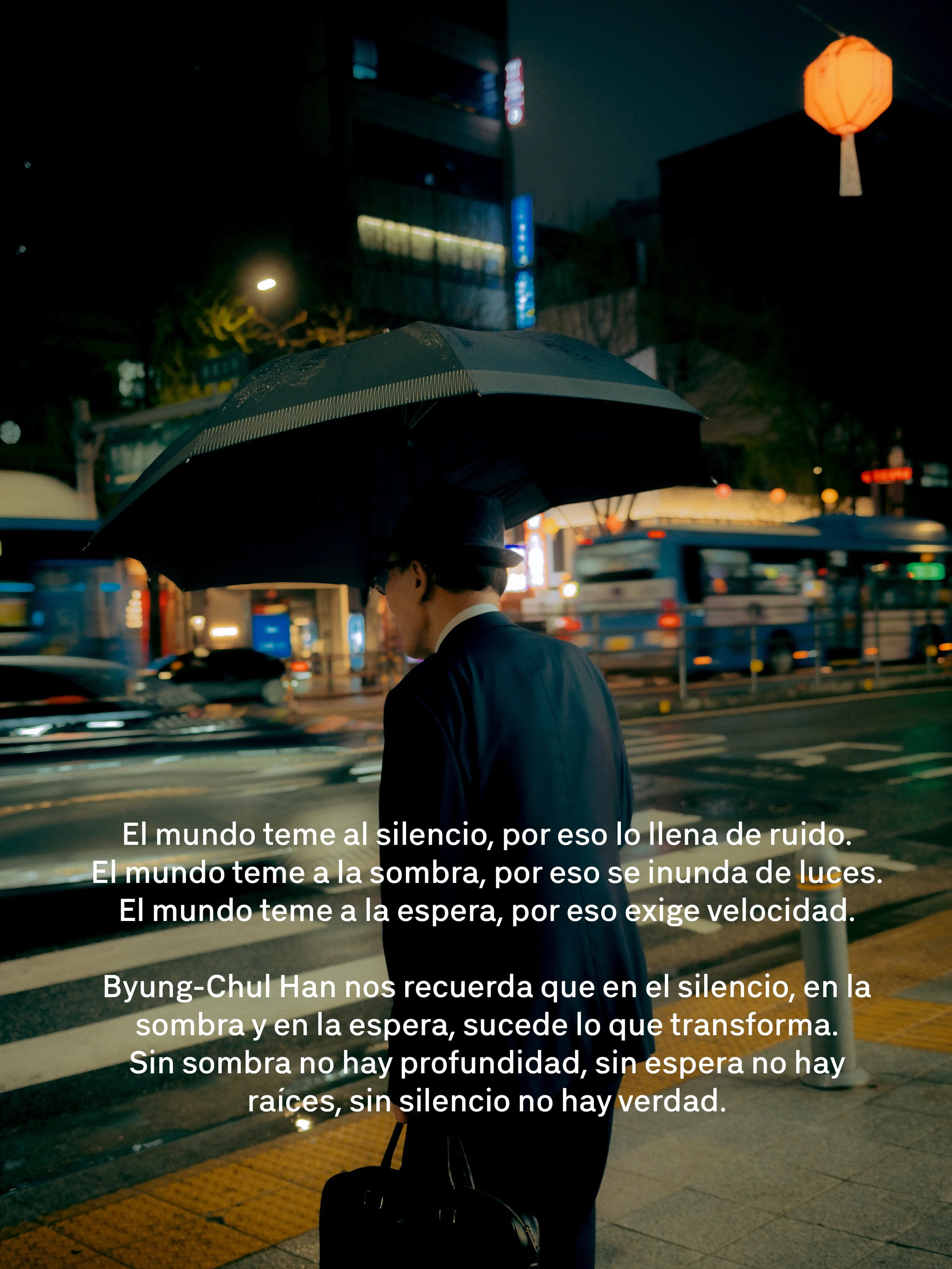 Una persona de espaldas con sombrero y maleta en una calle iluminada por luces y farolas en la noche. Texto superpuesto en la imagen en español que reflexiona sobre el silencio y la sombra.