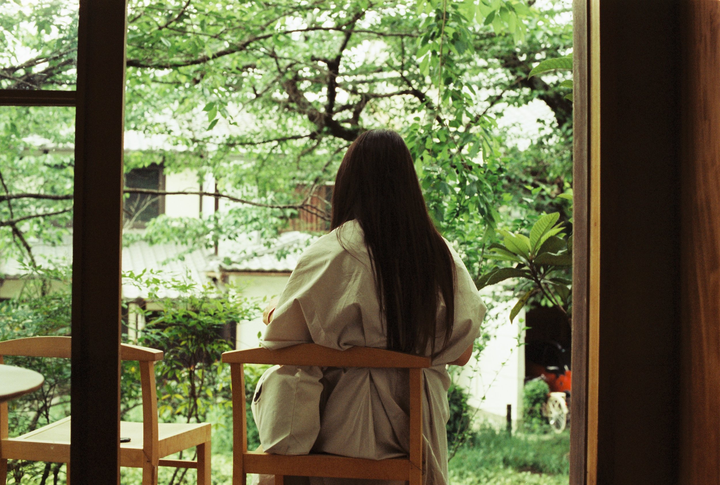 Mujer de espaldas sentada en una silla de madera mirándote por una ventana grande, rodeada de árboles verdes y vegetación en un ambiente natural y tranquilo.