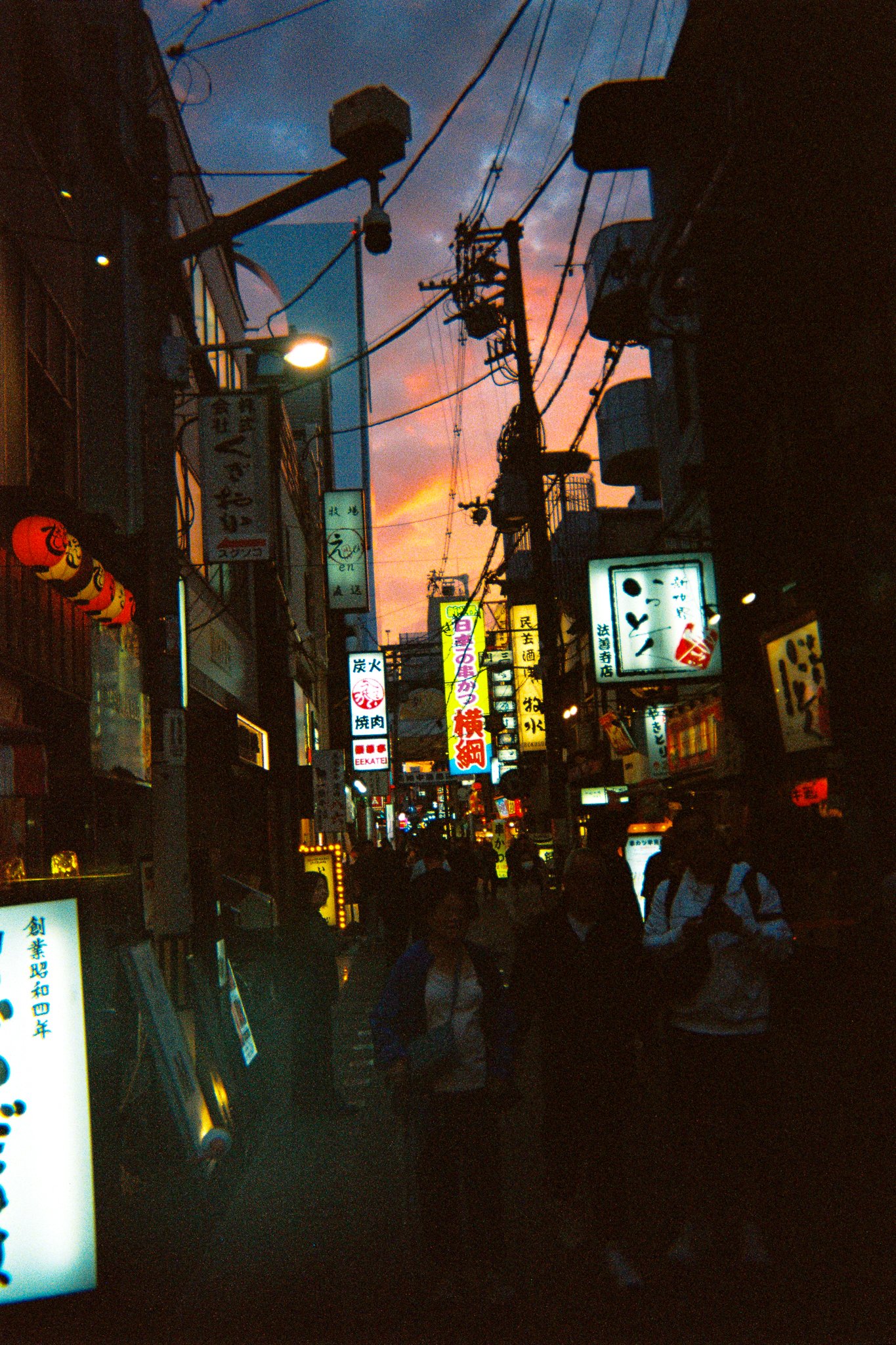 Escena nocturna en una calle de Japón con carteles iluminados en japonés, personas caminando y cables eléctricos en el cielo, con un atardecer colorido.