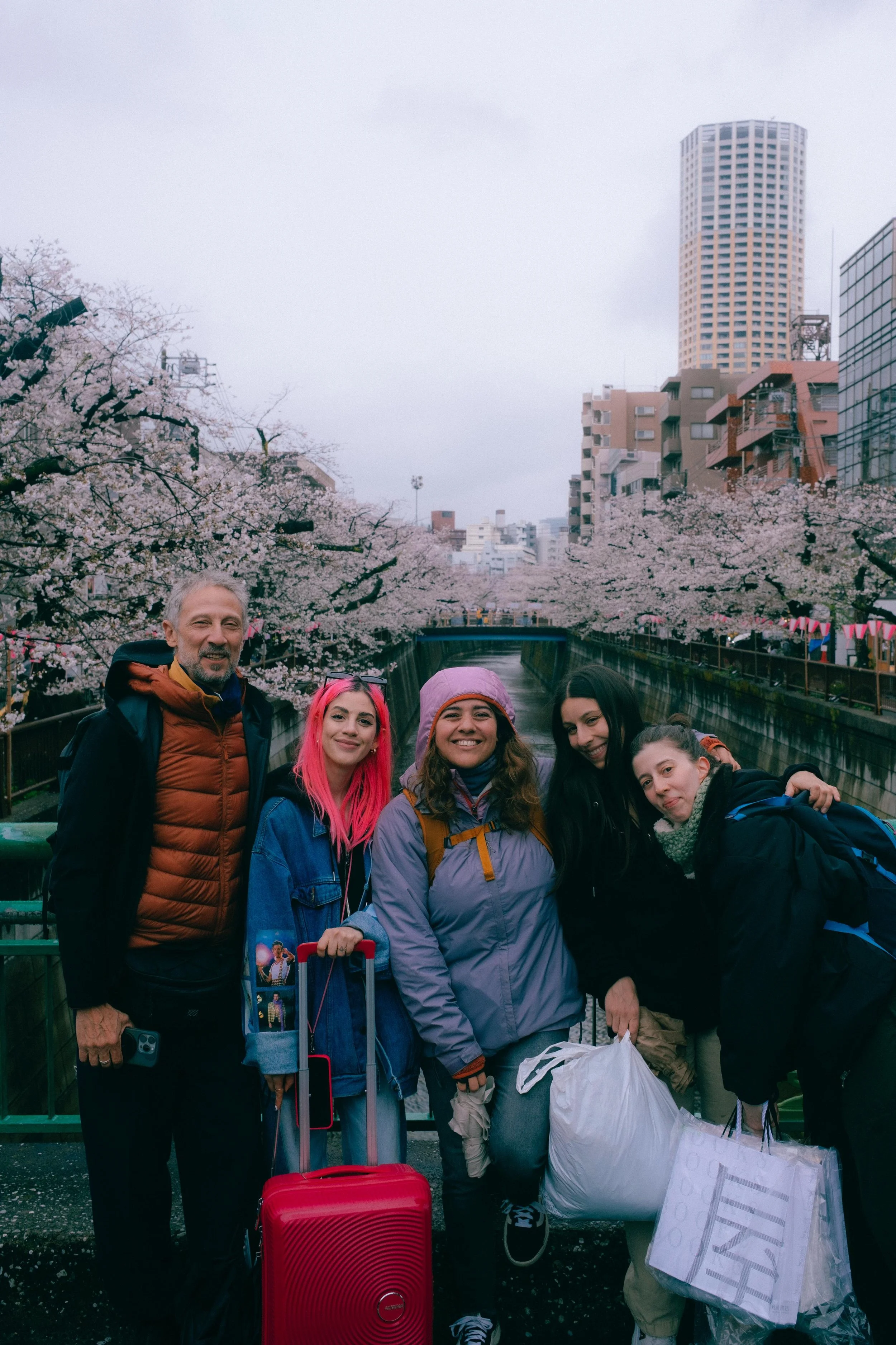 Grupo de cinco personas con mochilas y equipaje en un puente peatonal sobre un río con árboles en flor y edificios altos en ciudad. La escena es en un día nublado.