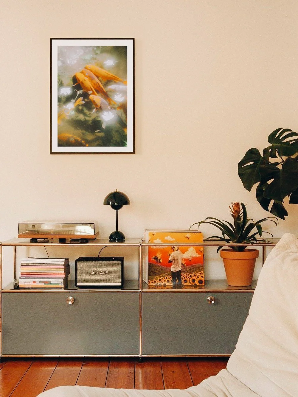 Pared beige con una pintura de peces enmarcada y un mueble con colecciones de discos, una lámpara, una bocina Marshall, una planta en maceta y un cuadro con una persona en un campo de girasoles.