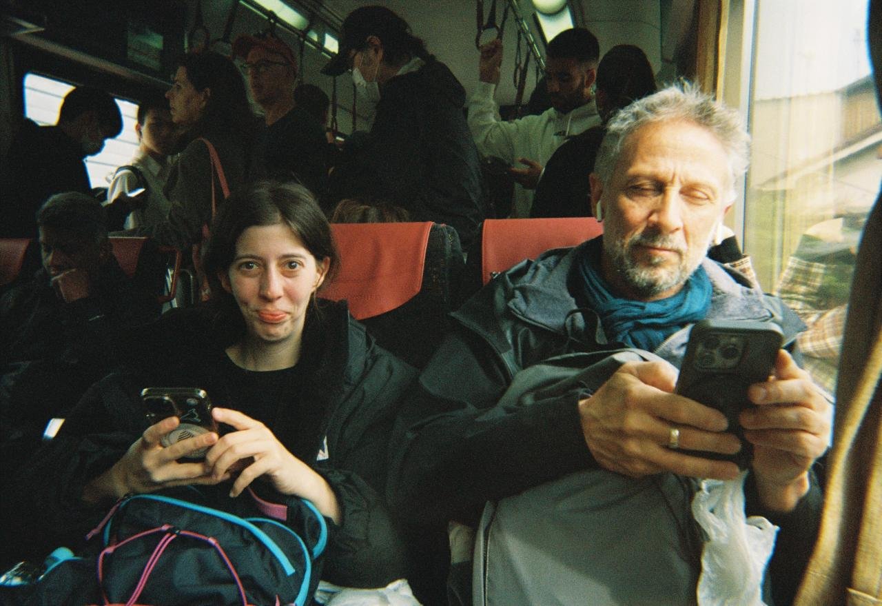 Una mujer joven y un hombre mayor sentados en un autobús, ambos usando teléfonos inteligentes, con muchas personas de pie y sentadas en el fondo.