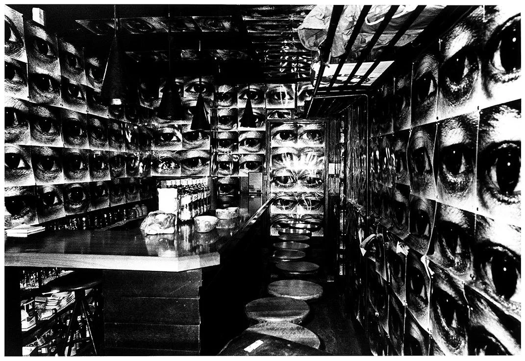 Interior de un bar con paredes decoradas con múltiples imágenes en blanco y negro de ojos humanos, mostrando una atmósfera artística y llamativa.