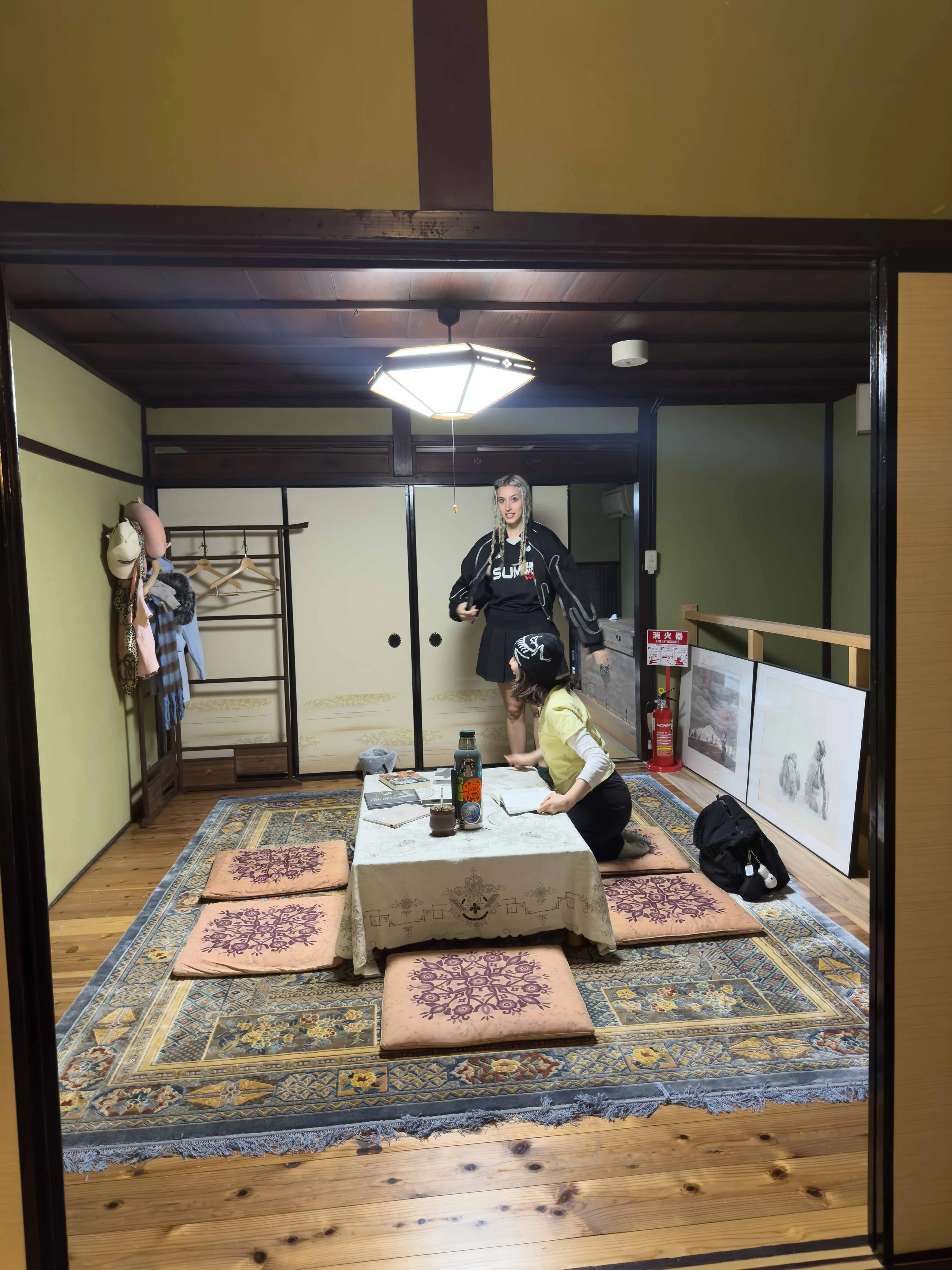 Una habitación tradicional japonesa con tatami y pufs, personas sentadas en el suelo, decorada con cuadros y objetos culturales.