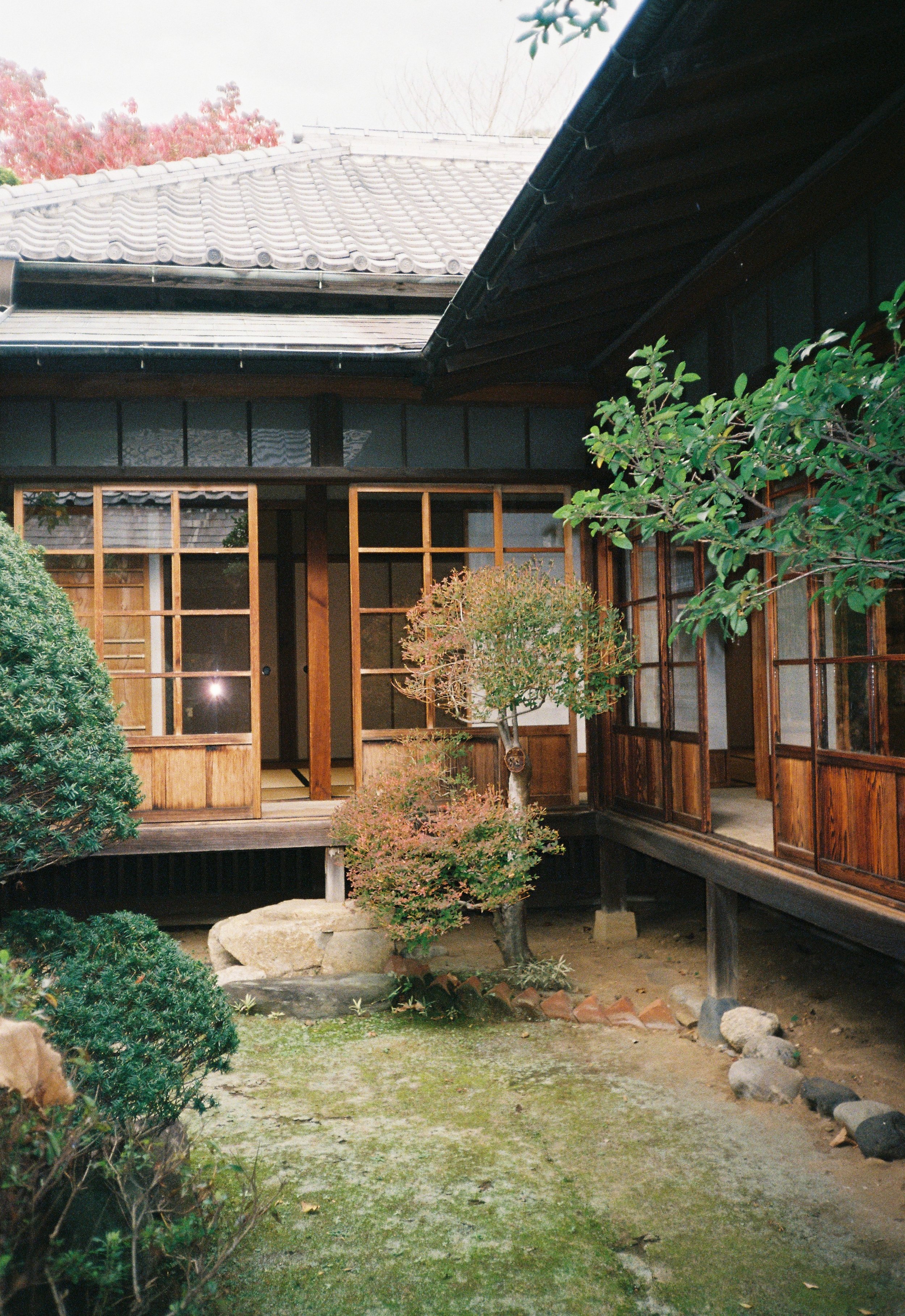 Casa tradicional de madera con ventanas grandes y puertas correderas, rodeada de un jardín con árboles y plantas, con un patio de tierra y piedras.