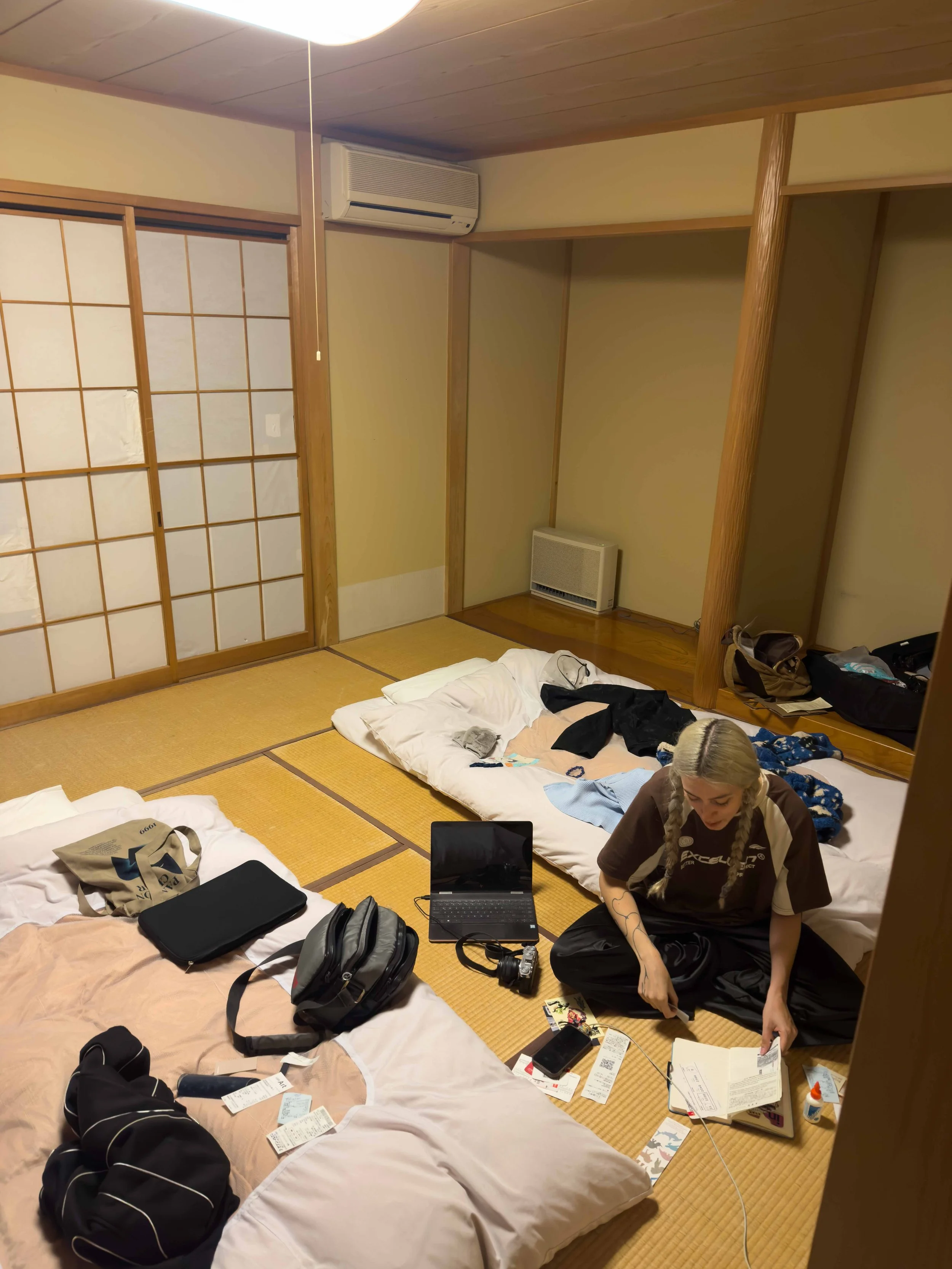 Una mujer joven en un cuarto tradicional japonés con tatamis, rodeada de maletas, libros y tecnología, mientras revisa una libreta.
